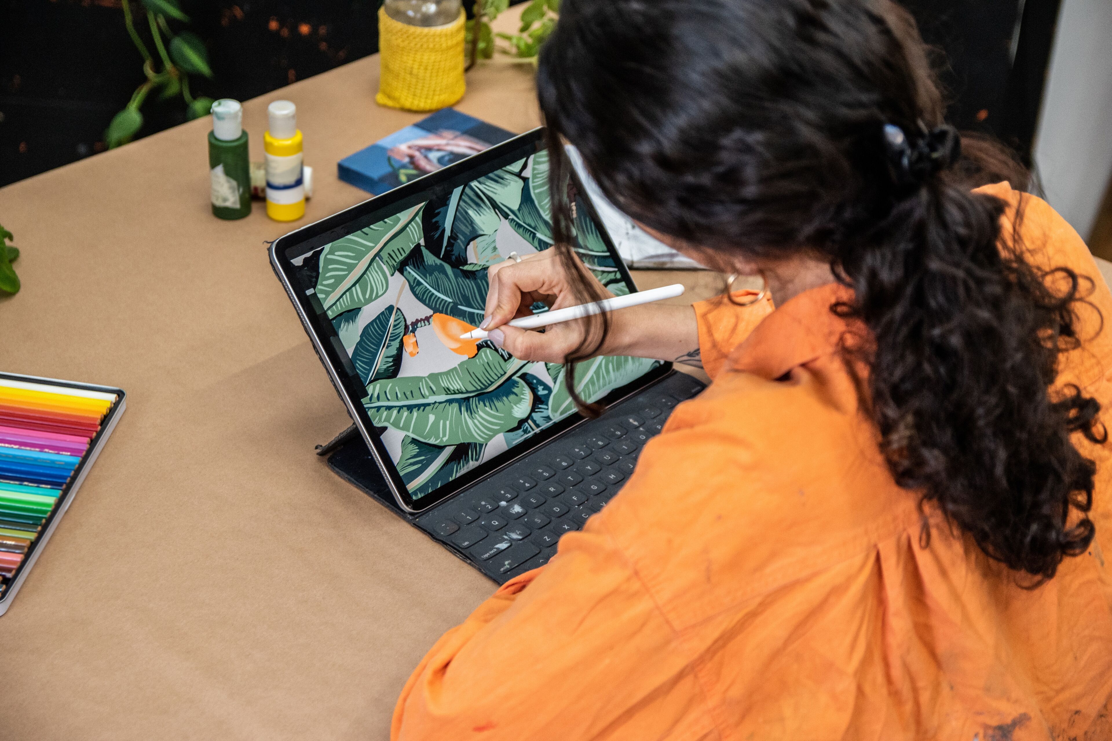Un artista con camisa naranja usa un lápiz óptico en una tableta para pintar digitalmente un diseño de hojas tropicales.