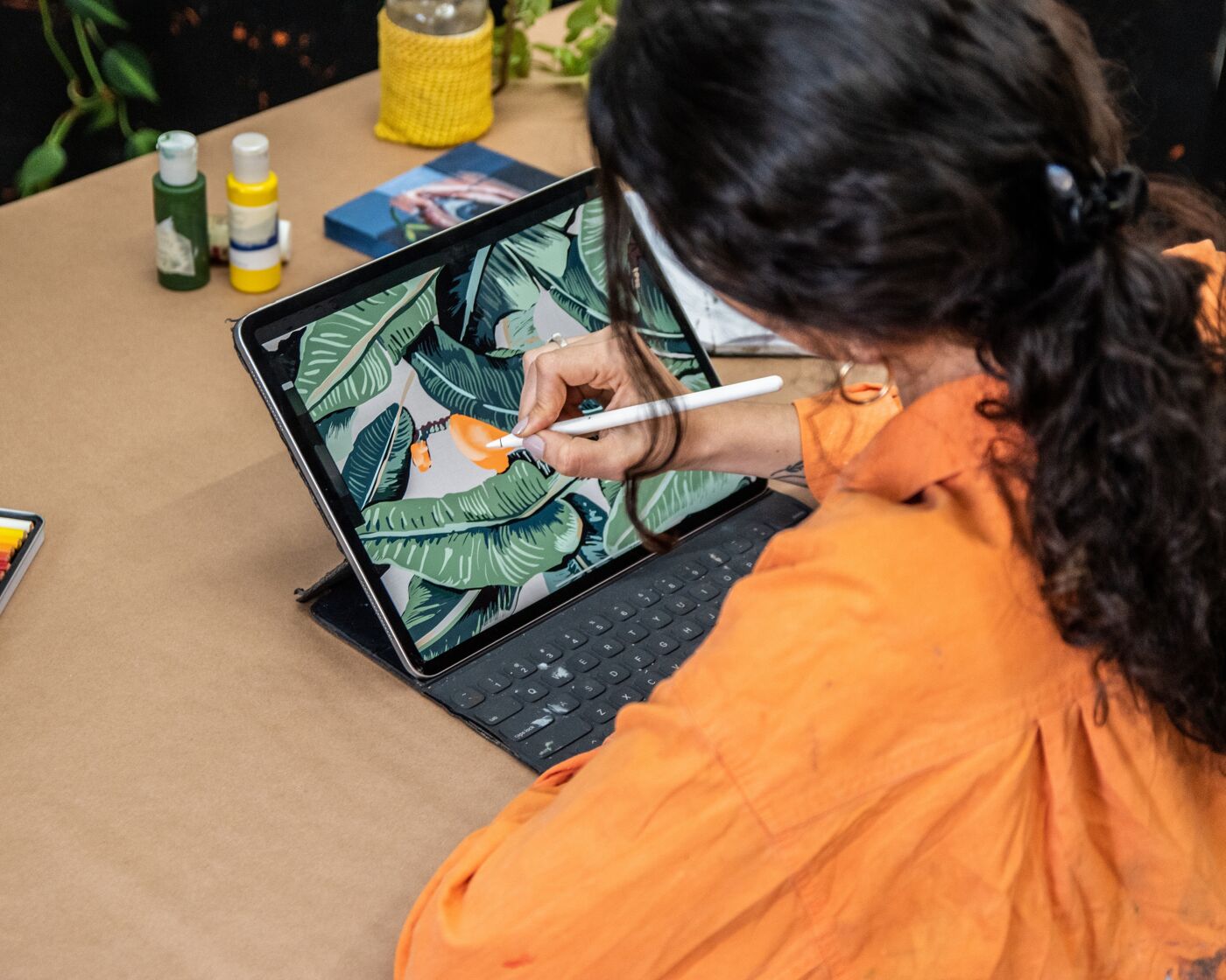 An artist in an orange shirt uses a stylus on a tablet to digitally paint a tropical leaf design.