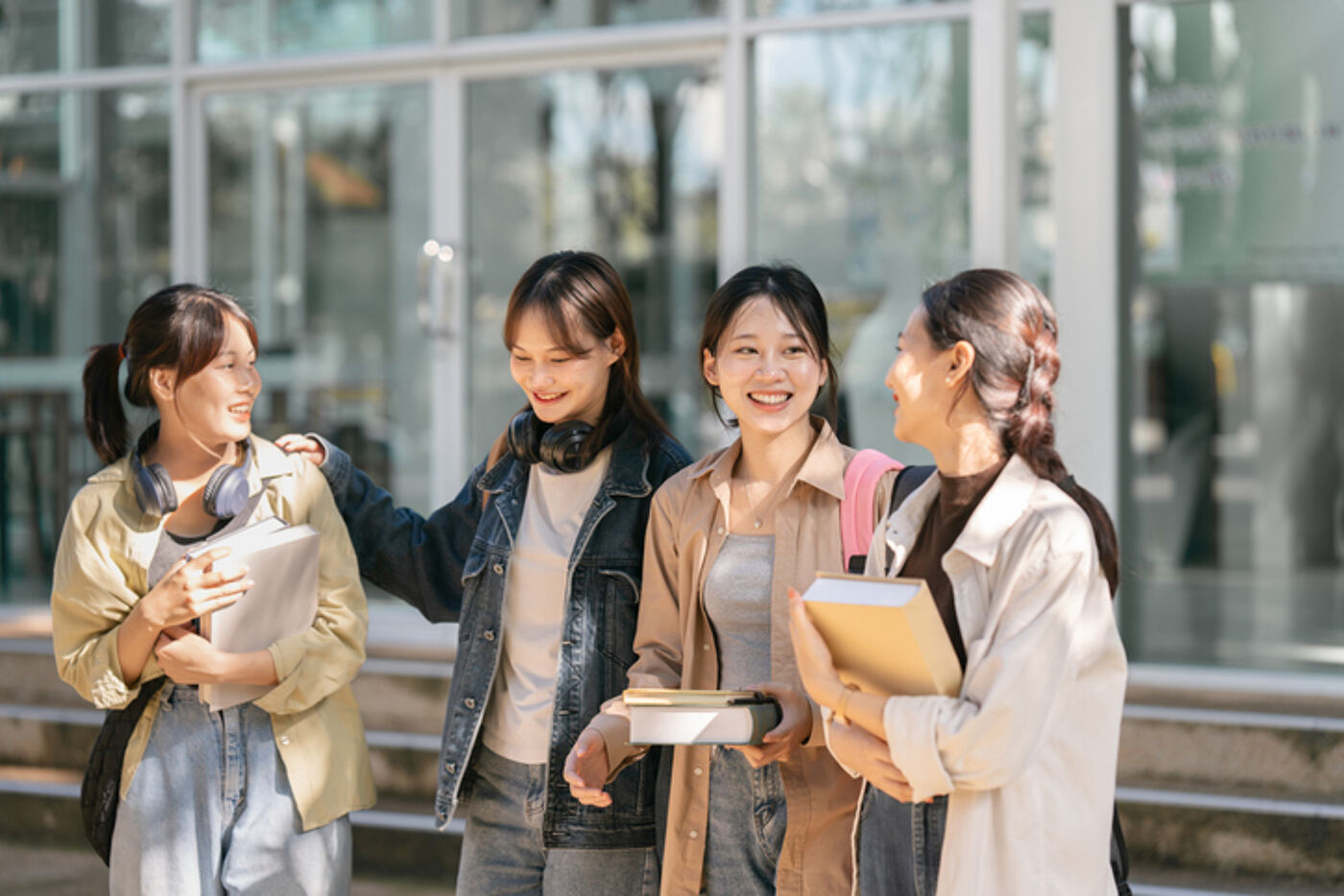 Four young women are standing outside a building, possibly a school or university. They are dressed casually and appear to be engaged in conversation, smiling and holding books or notebooks.