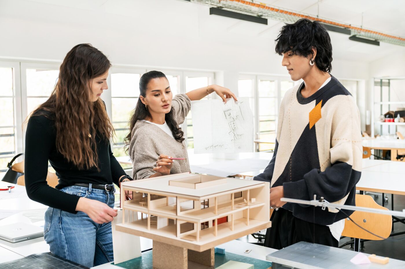 Three architecture students are discussing a wooden building model, pointing out details and reviewing papers for an academic project.