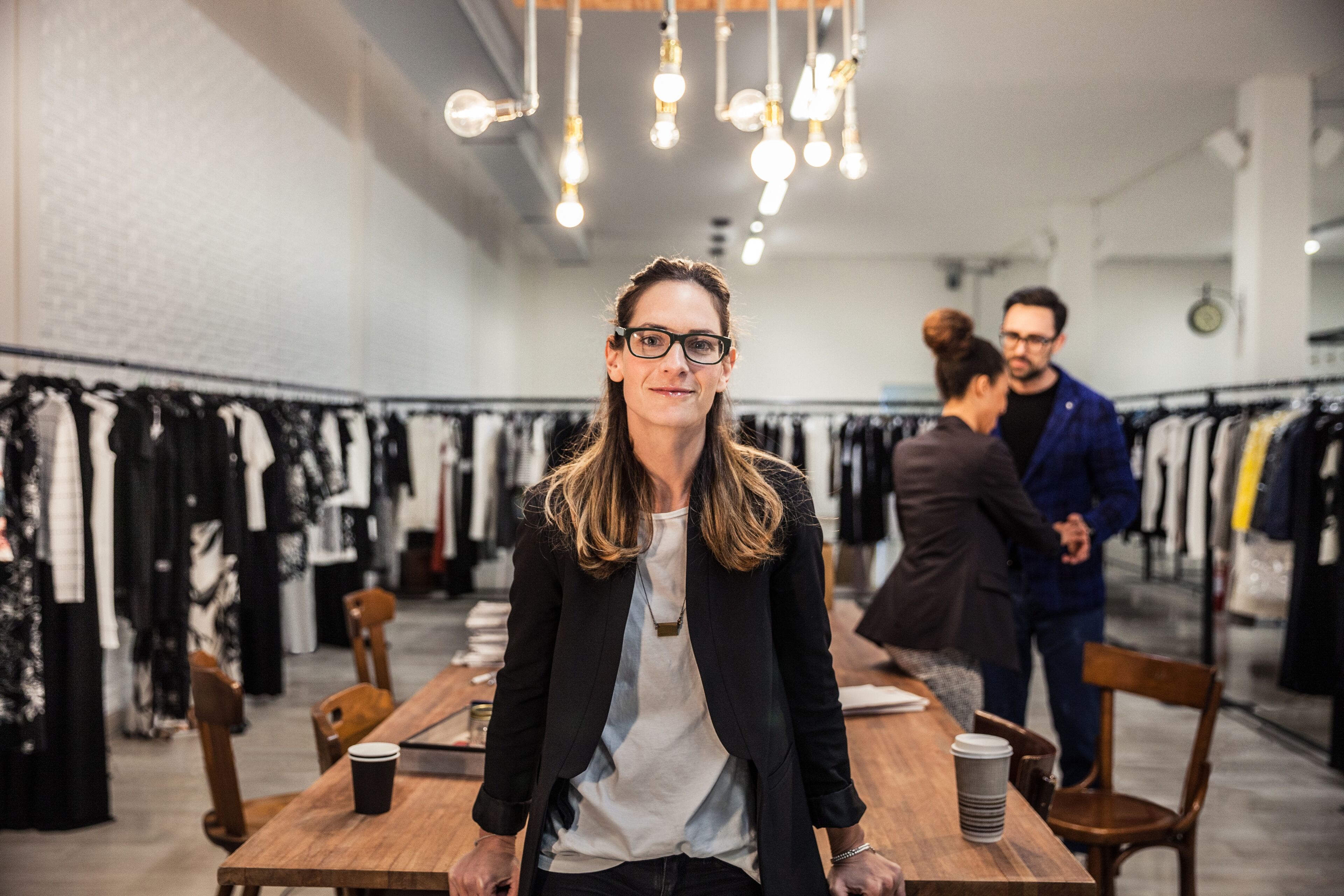 Una dona confiada amb ulleres somriu a la càmera en una boutique xic, amb penjadors de roba al fons.