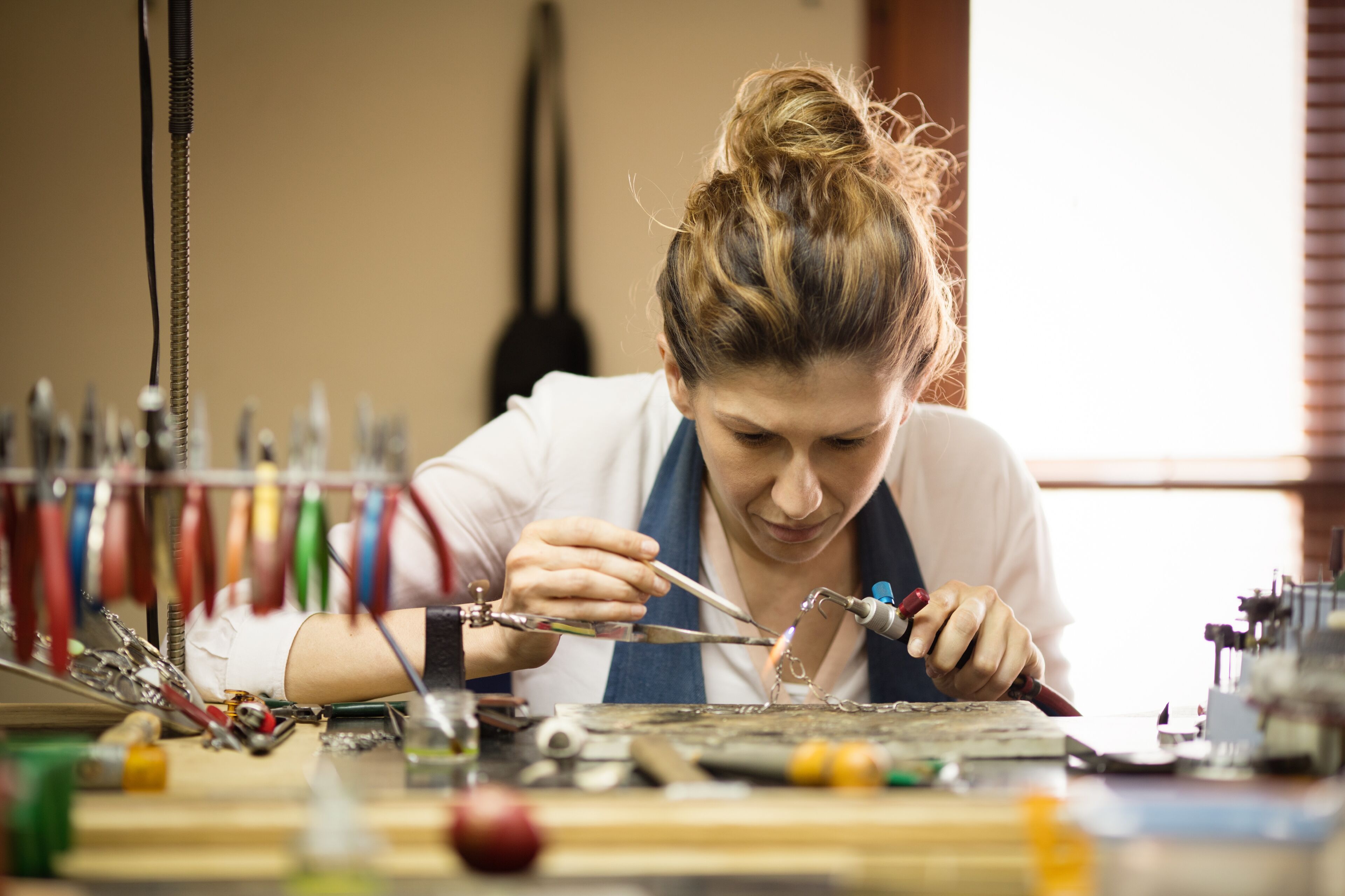 A focused female artisan soldering a piece of jewelry in a well-organized workshop.