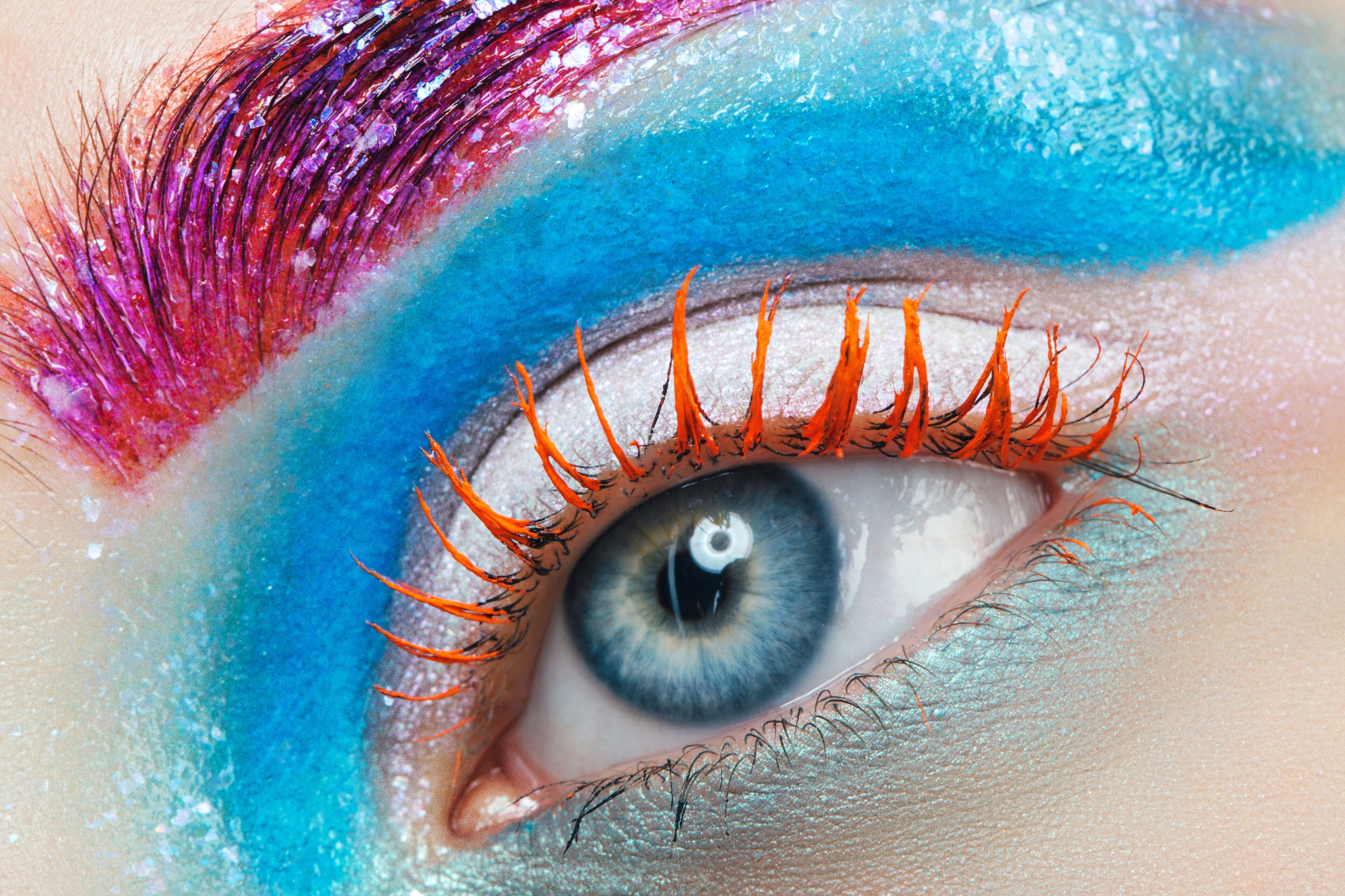 Close-up of a human eye with vibrant blue and pink eyeshadow, glitter, and orange mascara on eyelashes.