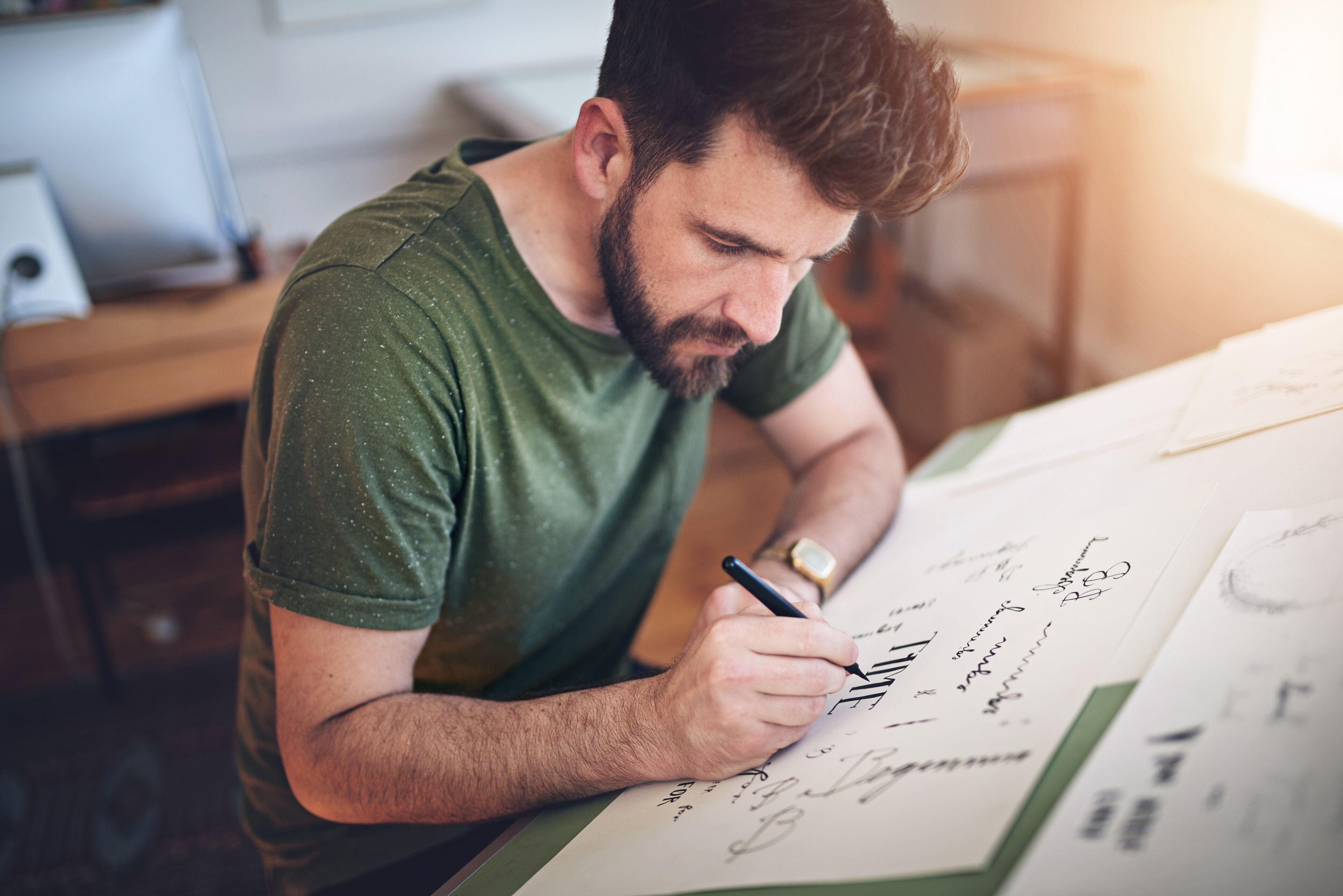 Un hombre adulto con barba está concentrado dibujando diseños en una hoja grande de papel, usando un bolígrafo en una habitación iluminada.