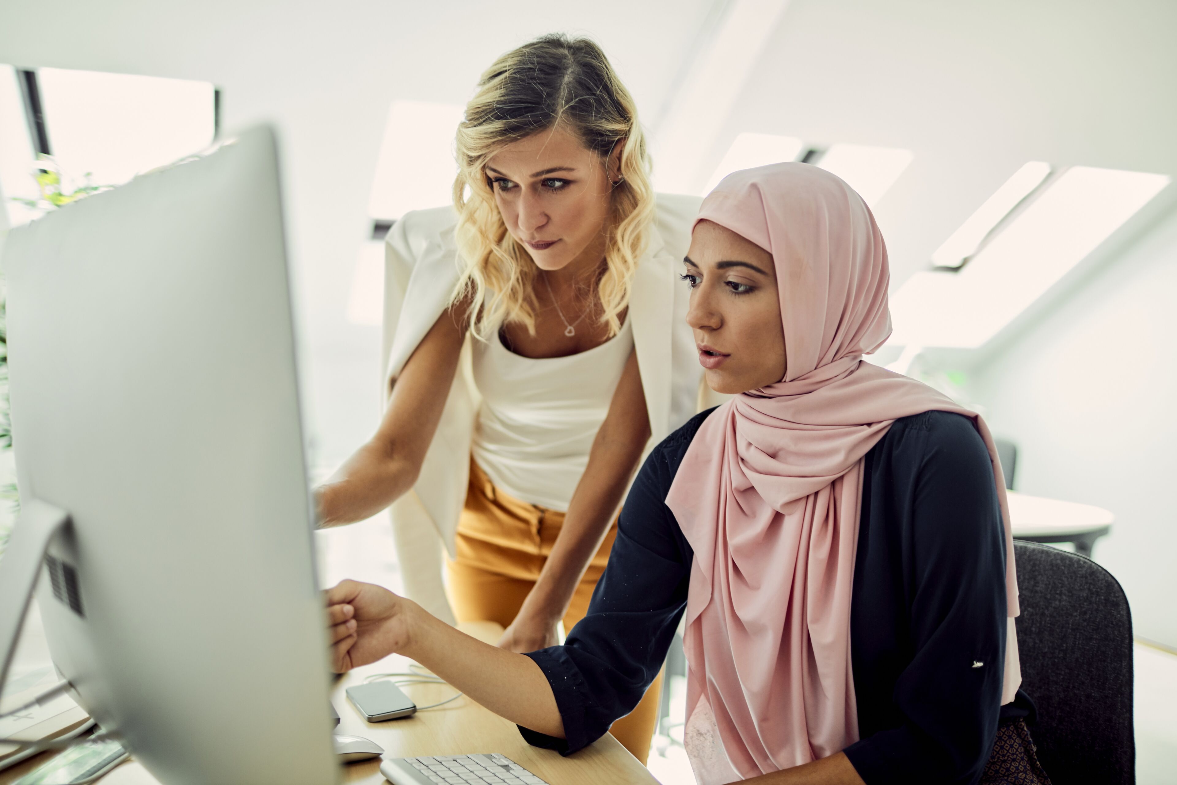 Deux femmes, une blonde et une autre portant un hijab, se concentrent sur un écran d'ordinateur, collaborant dans un environnement de bureau moderne.