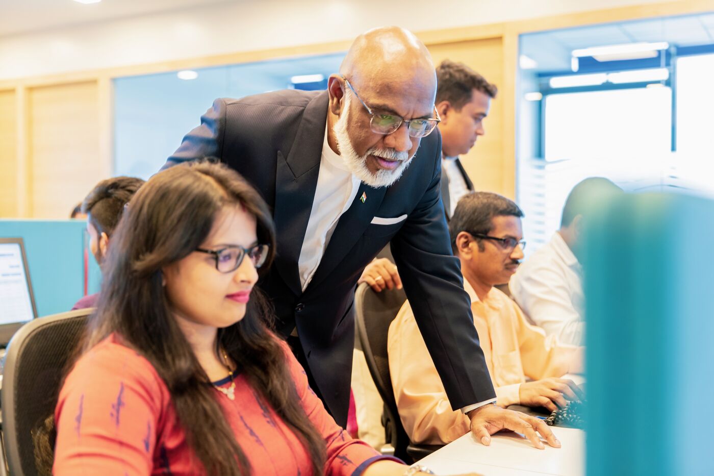 An elder male executive in a dark suit leans over to observe a younger female colleague at her computer in a busy office environment.