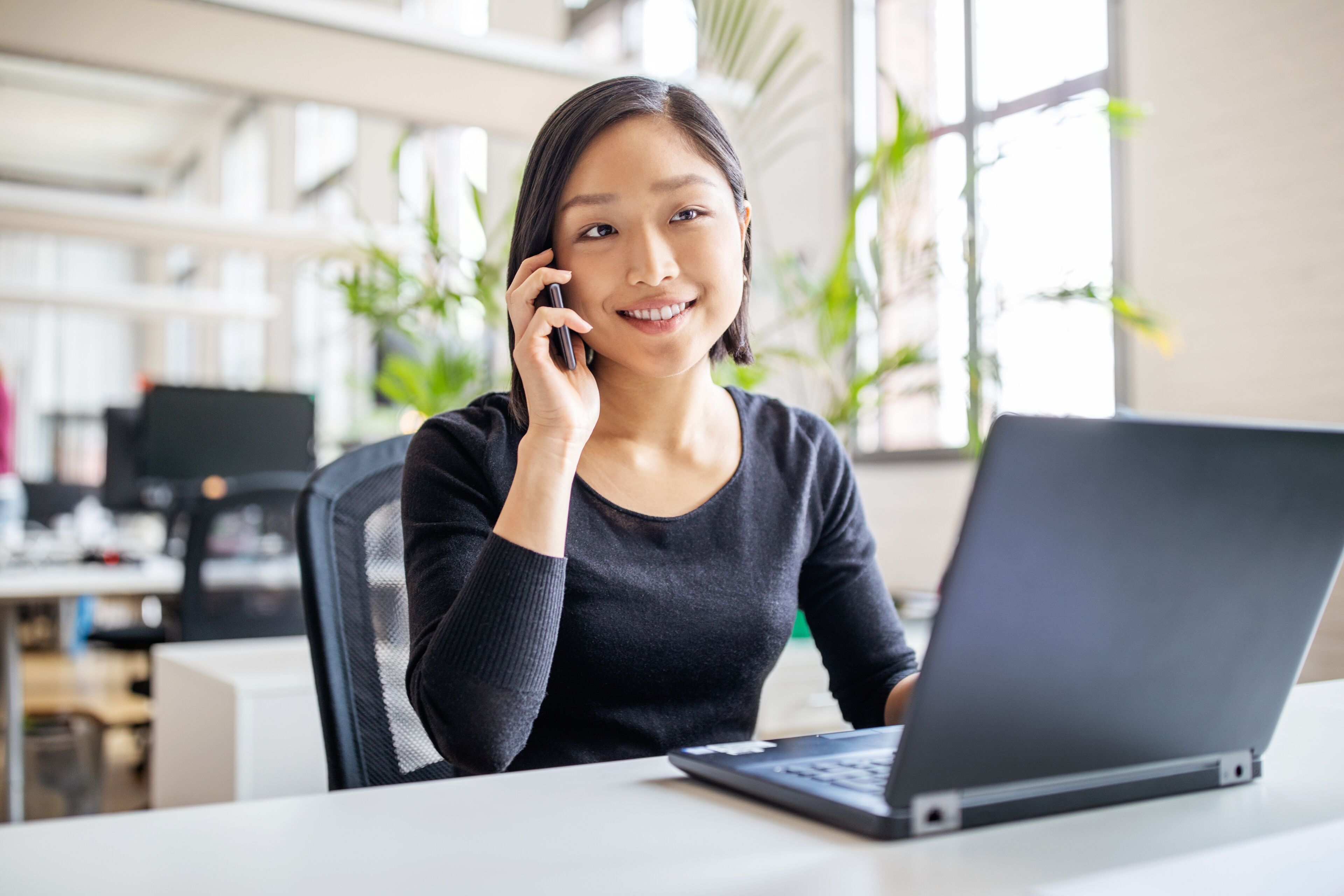 A young professional woman smiles while talking on the phone, seated at her desk with a laptop open in a bright office environment.