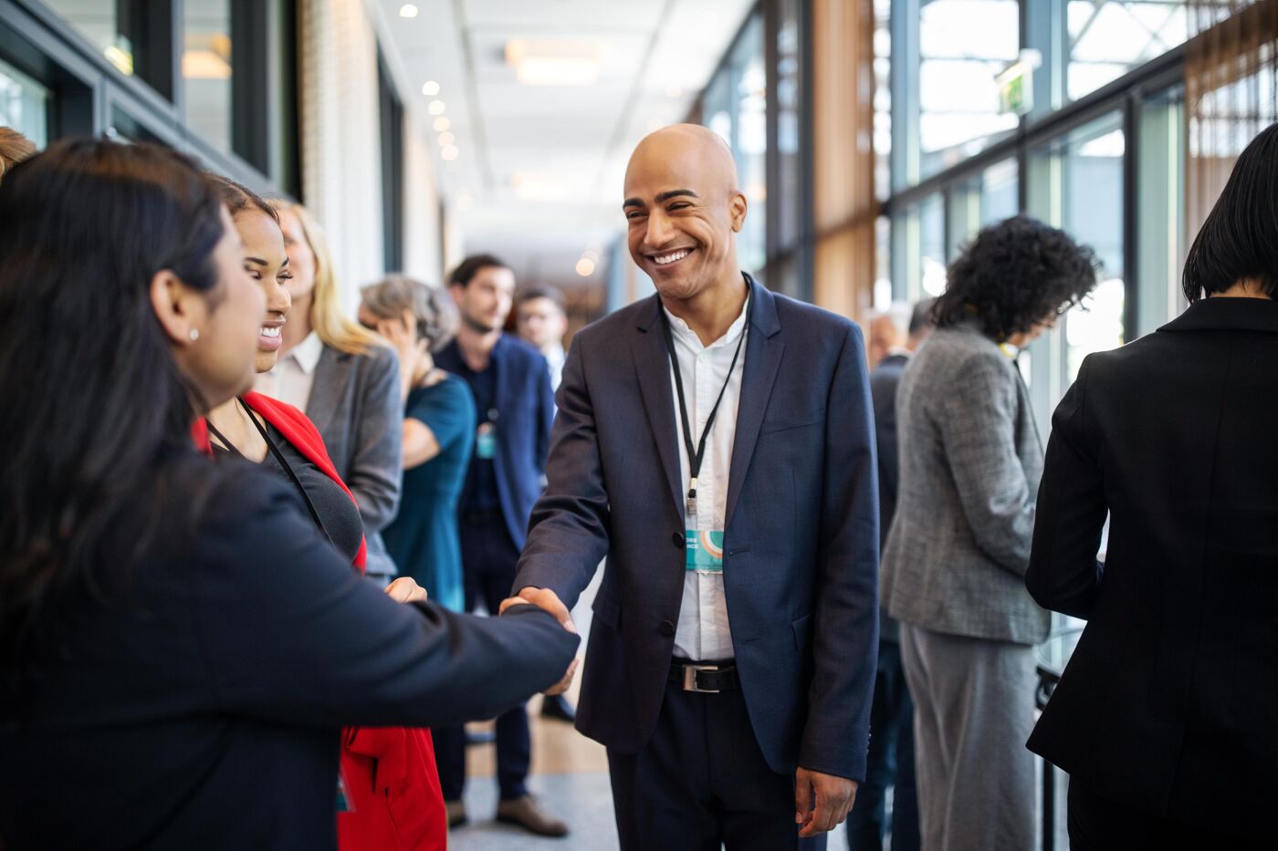 A man in a navy blue suit smiles as he shakes hands with a woman during a networking event. Other participants, engaged in conversations, are visible in the background. The event is held in a modern, well-lit venue with large windows and a professional atmosphere.