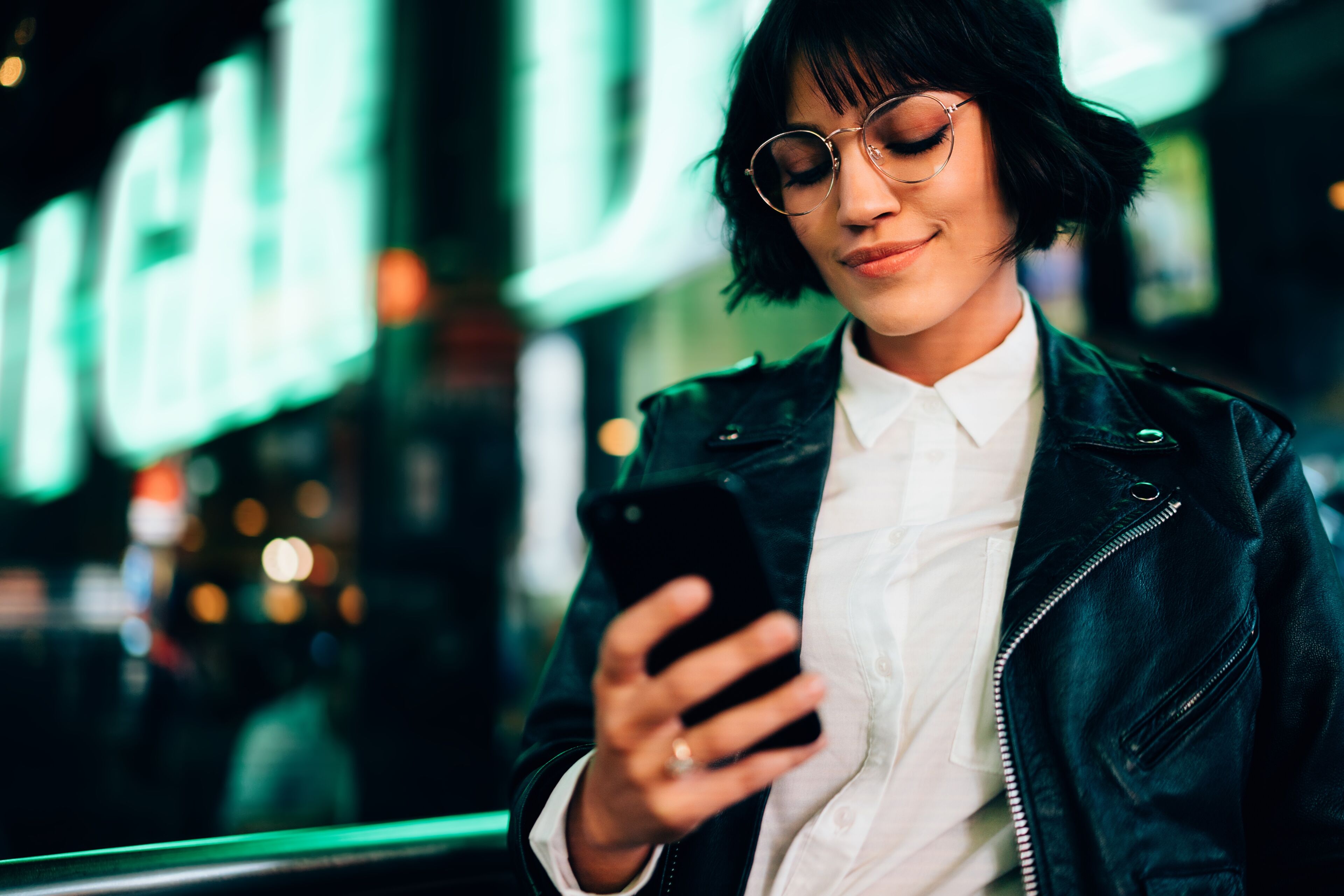 A young woman in a leather jacket and round glasses is focused on her smartphone, with bright city lights blurred in the background.