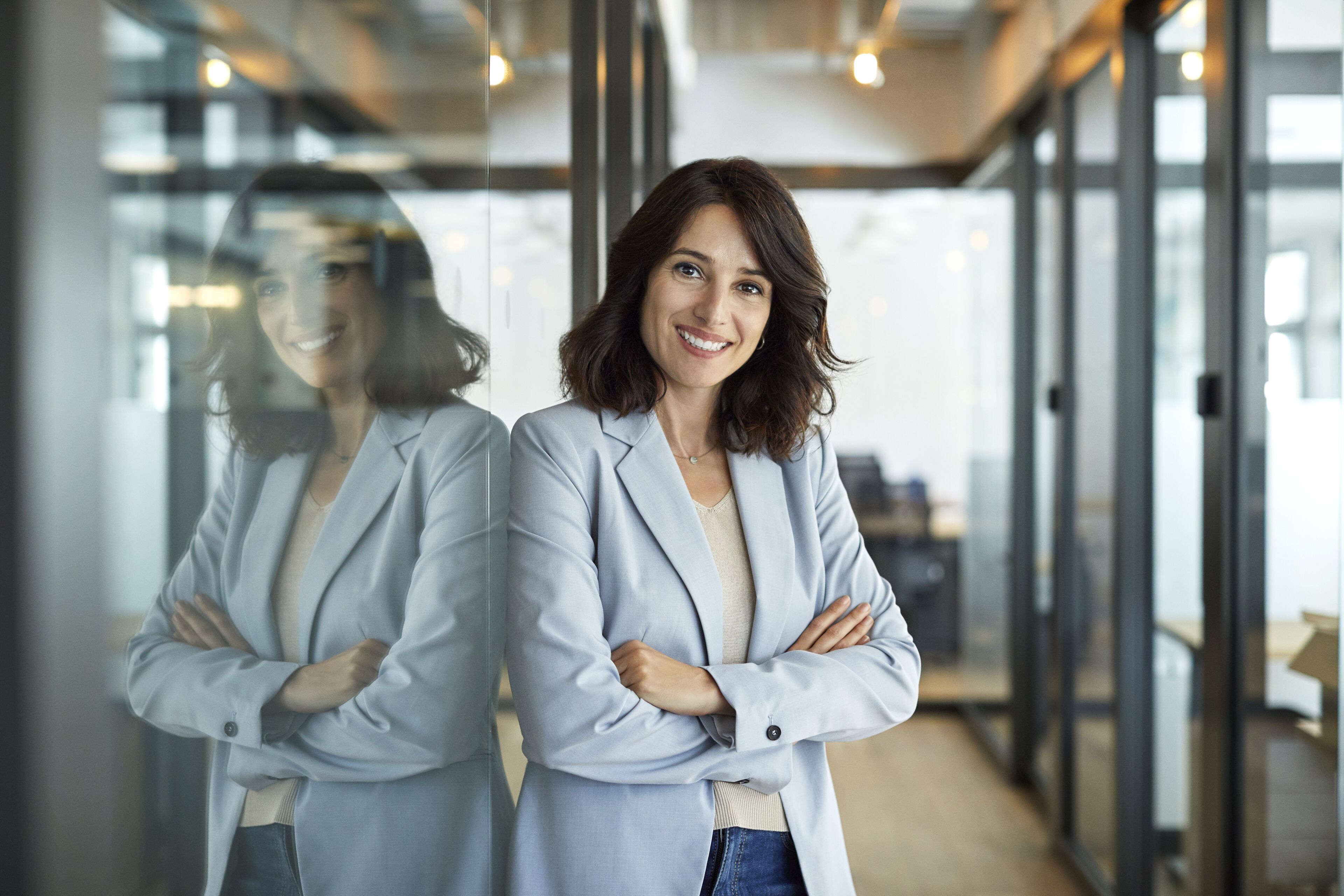 A professional woman in a light grey blazer stands with crossed arms, smiling in a modern office setting, reflecting confidence and approachability.