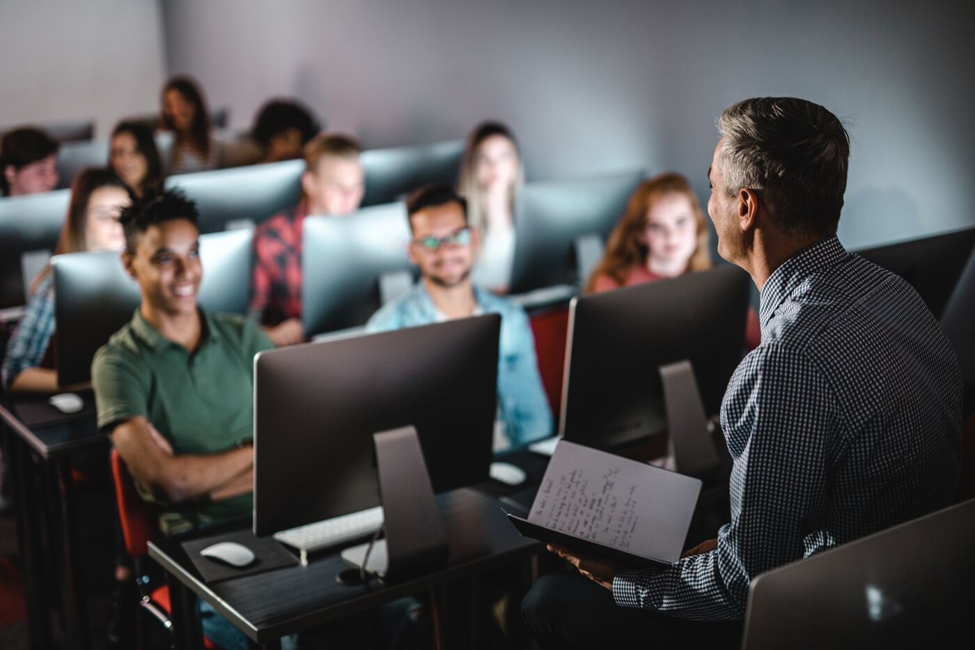 A professor engages with diverse students using laptops in a dimly lit lecture hall.