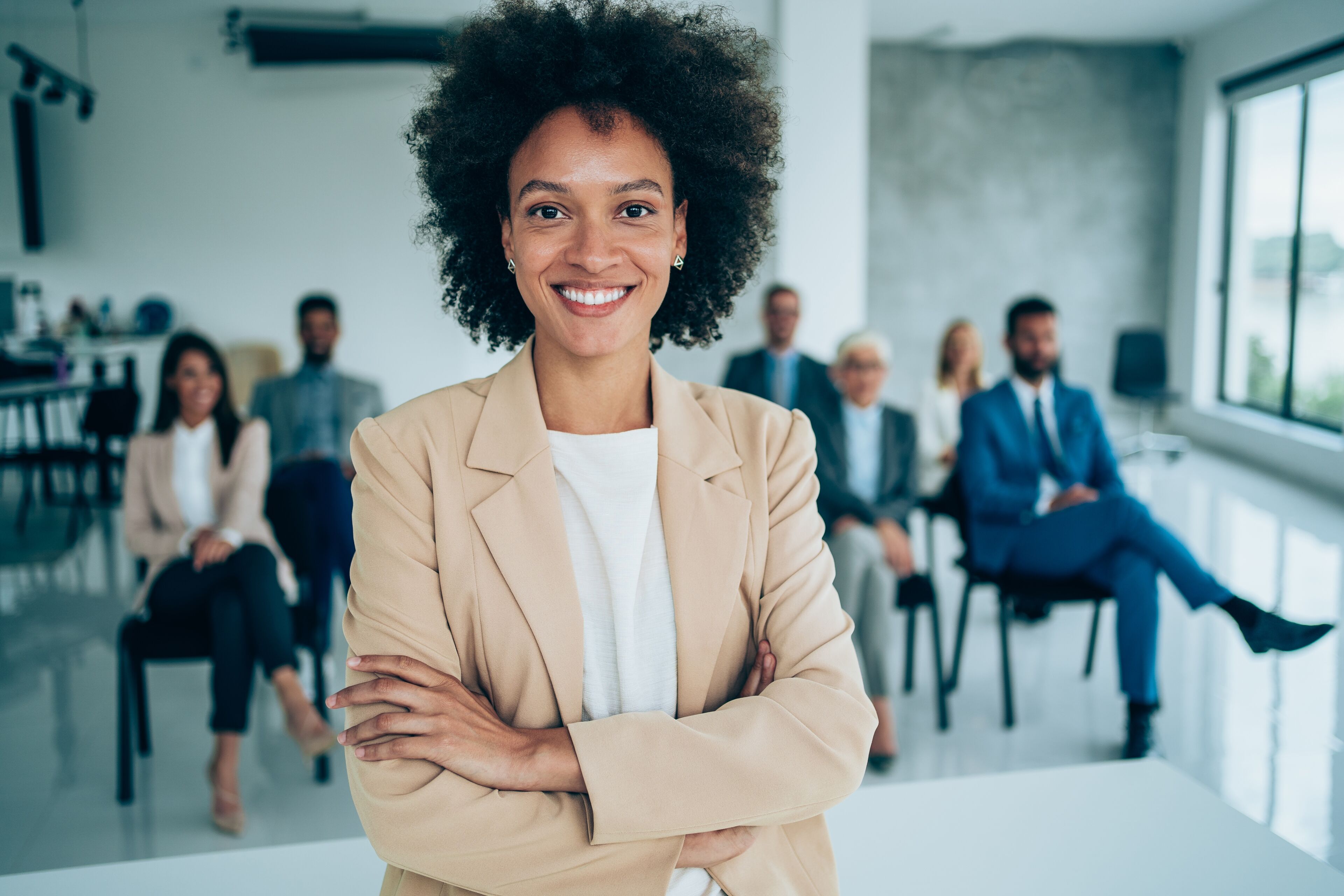 Una mujer afroamericana con pelo rizado se sitúa con confianza al frente, con los brazos cruzados, y un equipo de profesionales sentados detrás de ella en una oficina moderna.