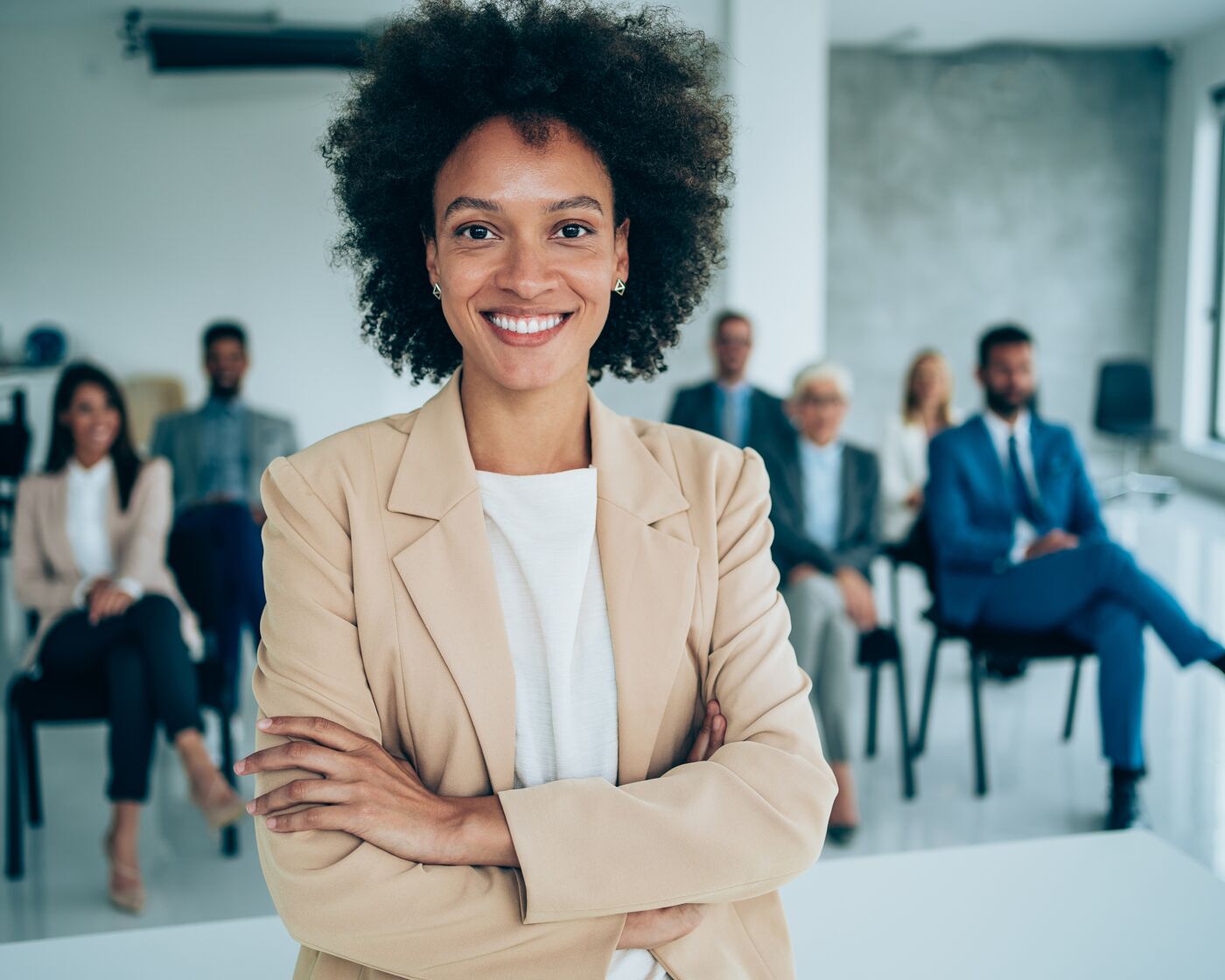 A poised African-American woman with curly hair stands confidently at the forefront, arms crossed, with a team of professionals seated behind her in a modern office setting.