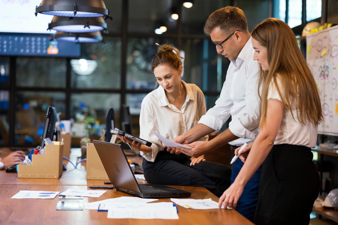 Three colleagues discuss documents in front of a laptop in a modern office setting, featuring industrial ceiling lights and a collaborative work atmosphere.