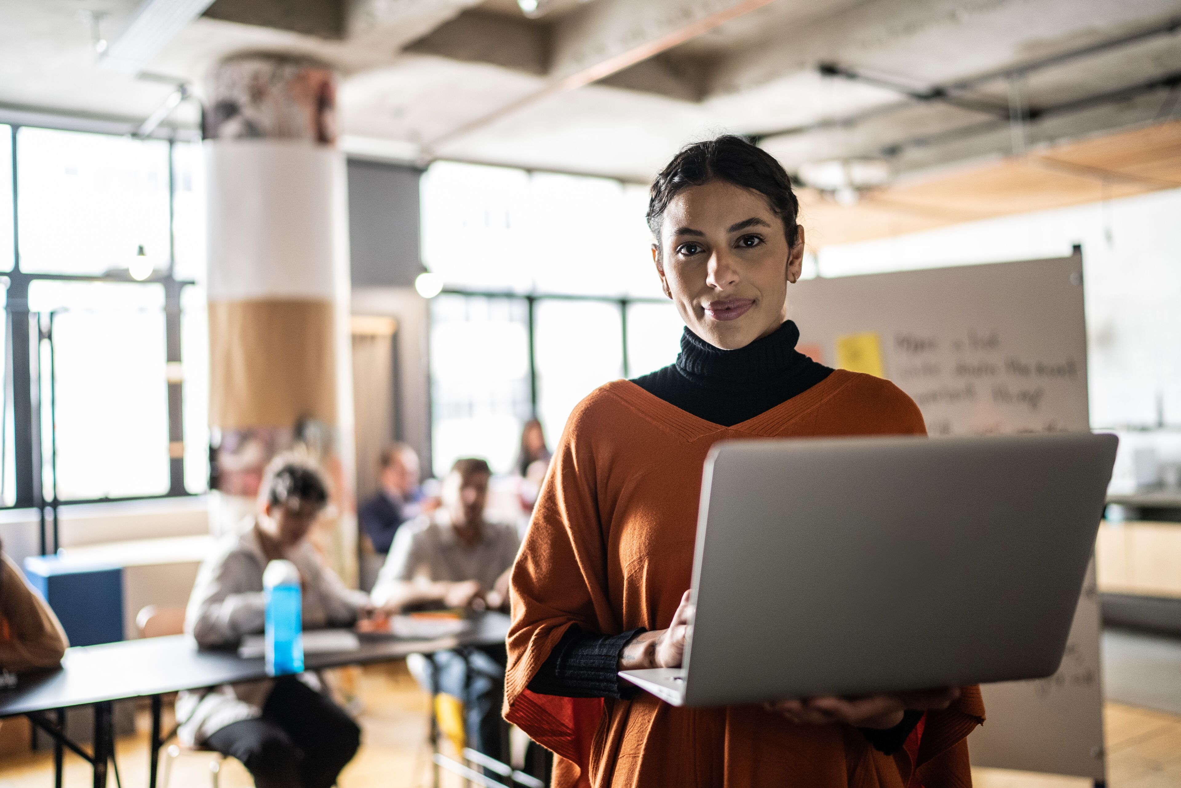 A focused female teacher in an orange sweater holds a laptop, standing in a classroom with students in the background, exemplifying active learning and leadership.