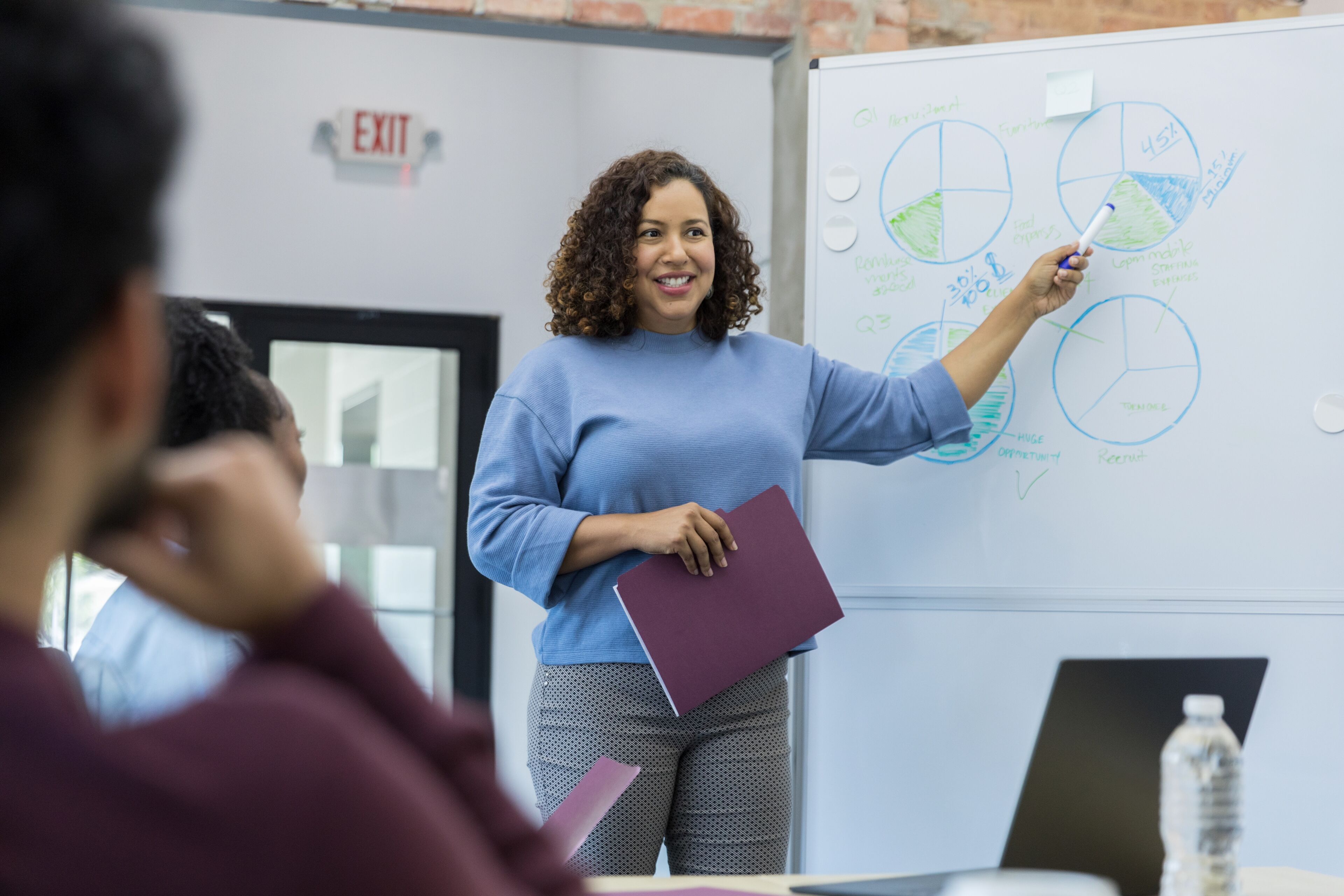 A professional woman presenting pie charts and data analysis on a whiteboard in a modern office, engaging with her colleagues.