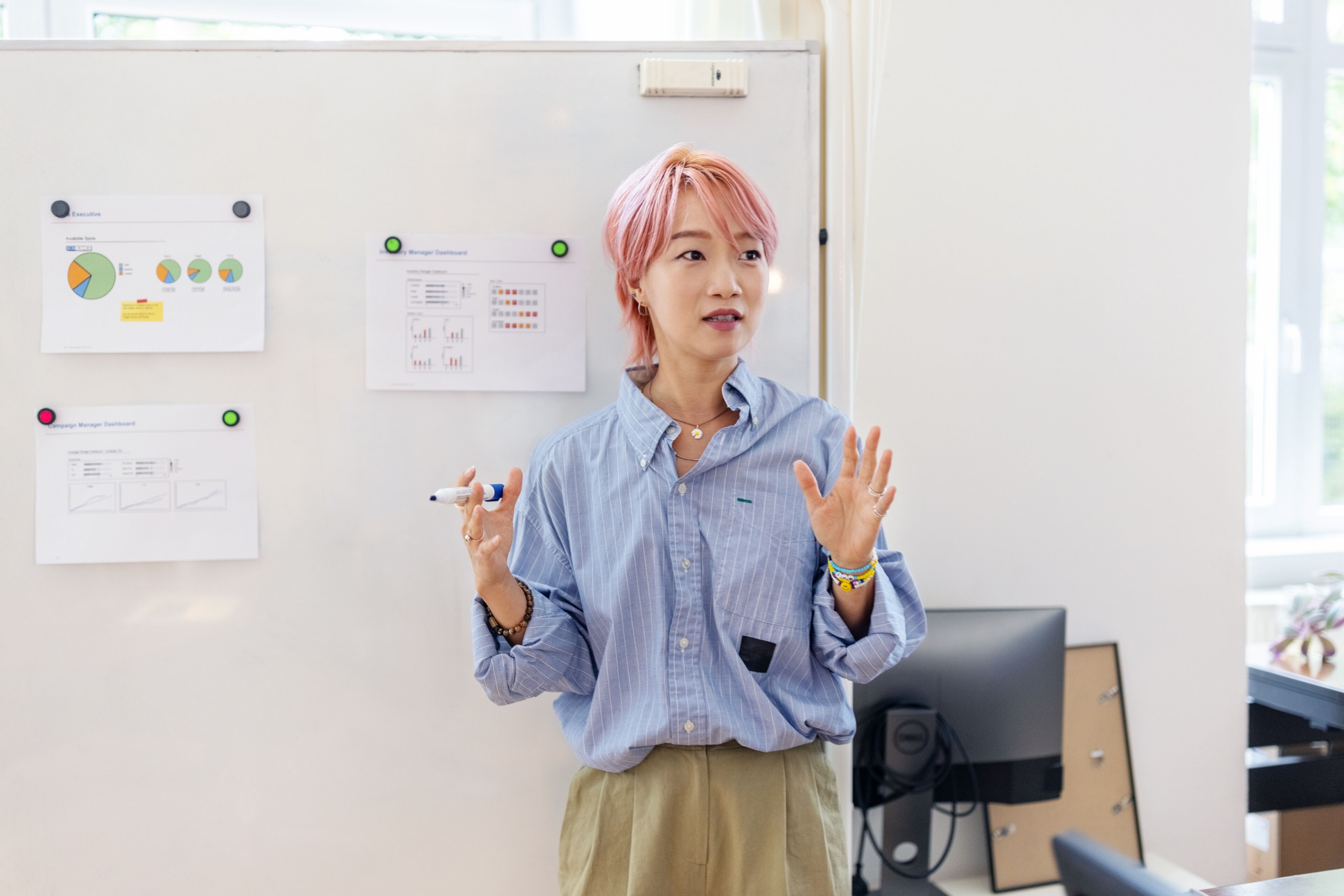A confident individual with pink hair delivers a presentation in a bright office, gesturing towards charts on a whiteboard.