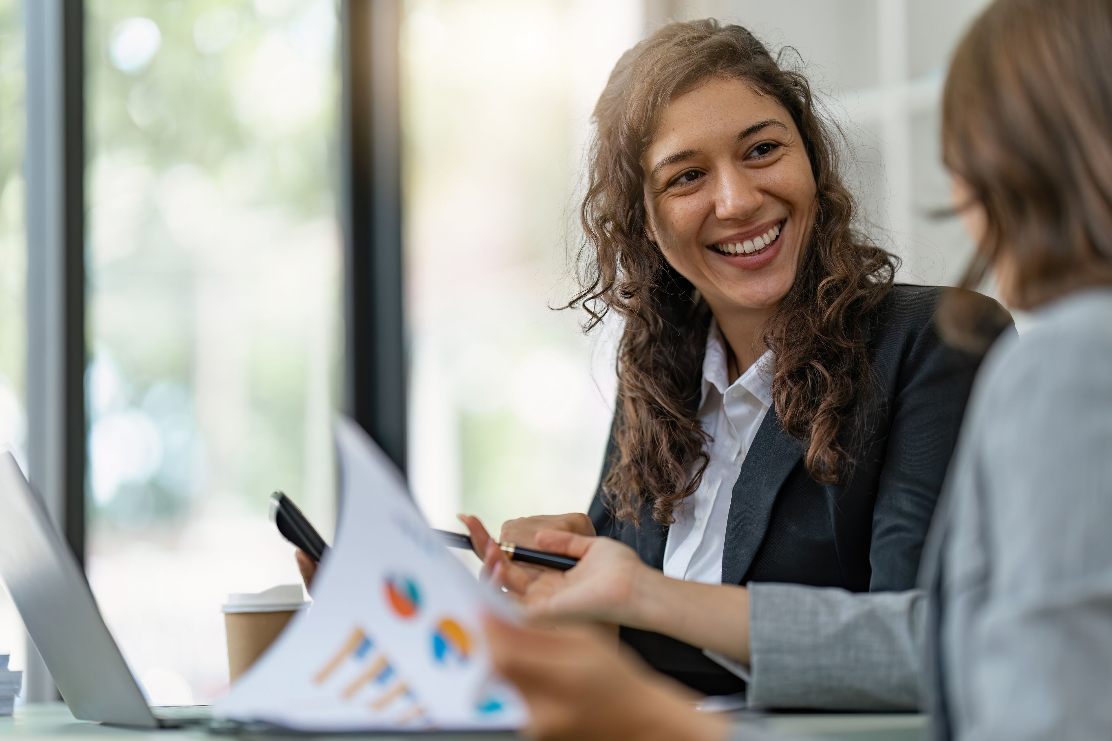 A cheerful businesswoman engages in a lively discussion with a colleague, with laptops and documents on the table, in a well-lit office.