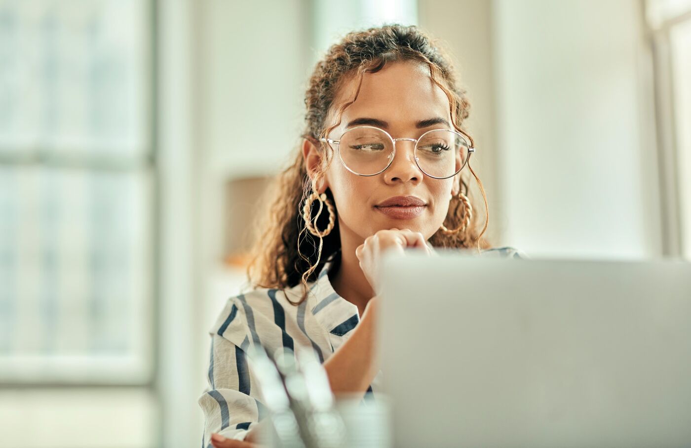 A young woman with glasses and large hoop earrings intently looks at a laptop screen, resting her chin on her hand.