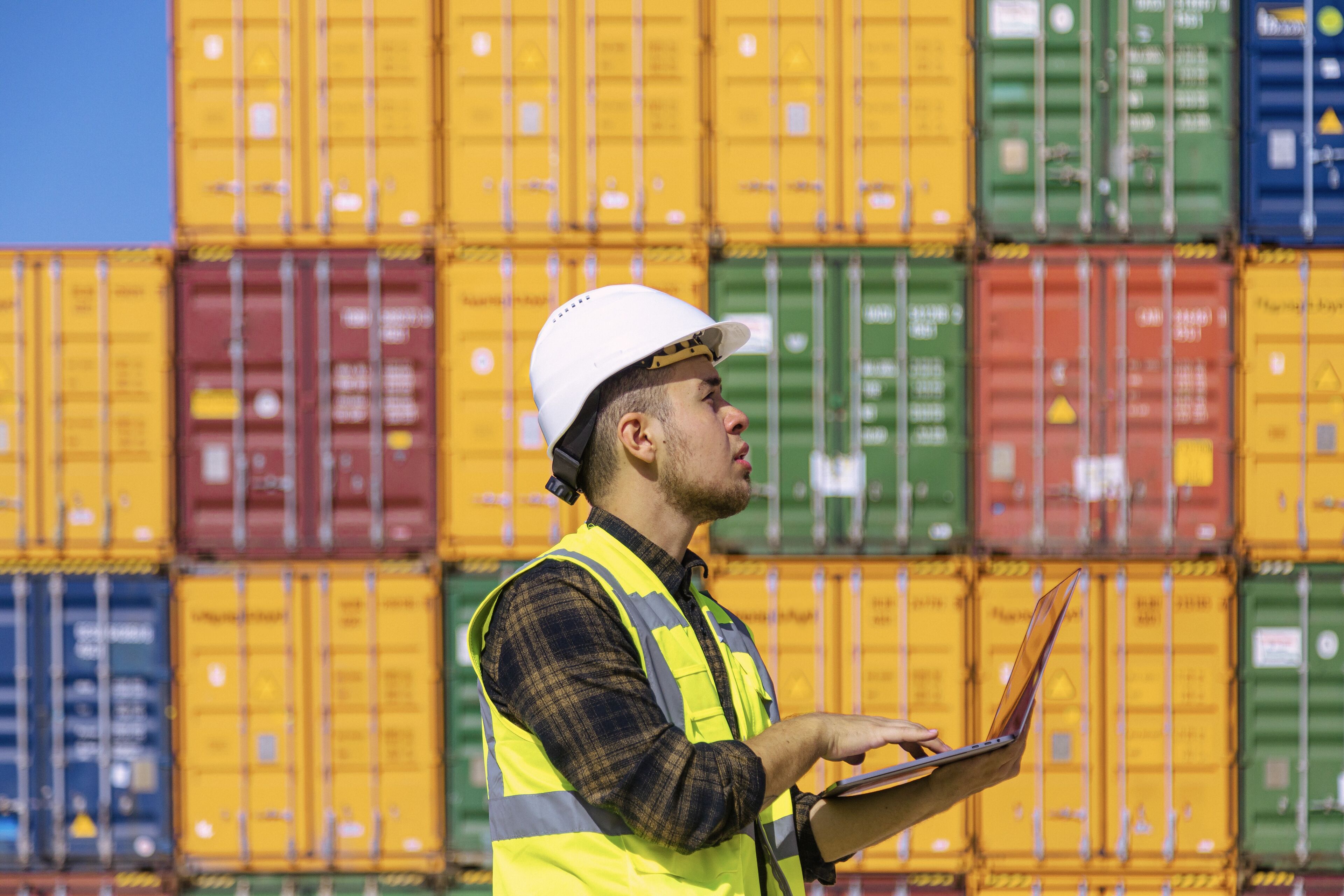 A man in a safety vest and helmet inspects paperwork with cargo containers in the background.