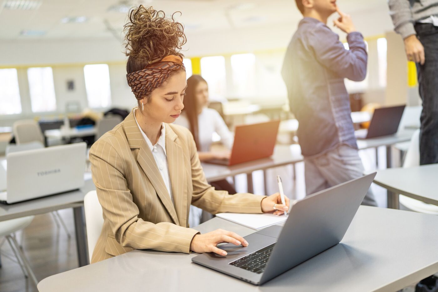 A focused businesswoman works on her laptop in a bright office space with colleagues in the background.