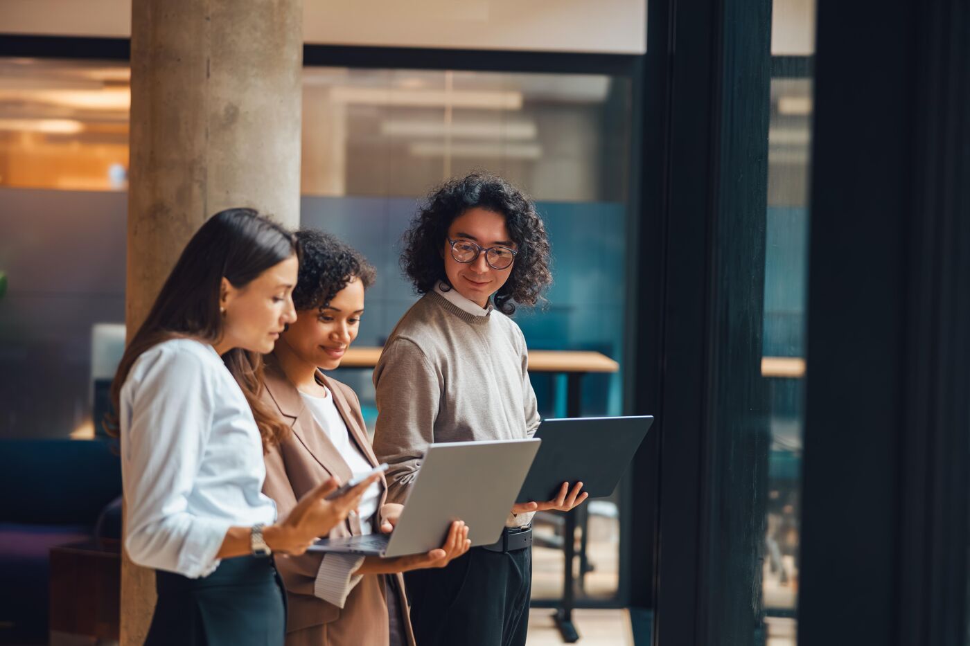 Three diverse women are gathered around a laptop, discussing work in a modern, naturally lit office setting.