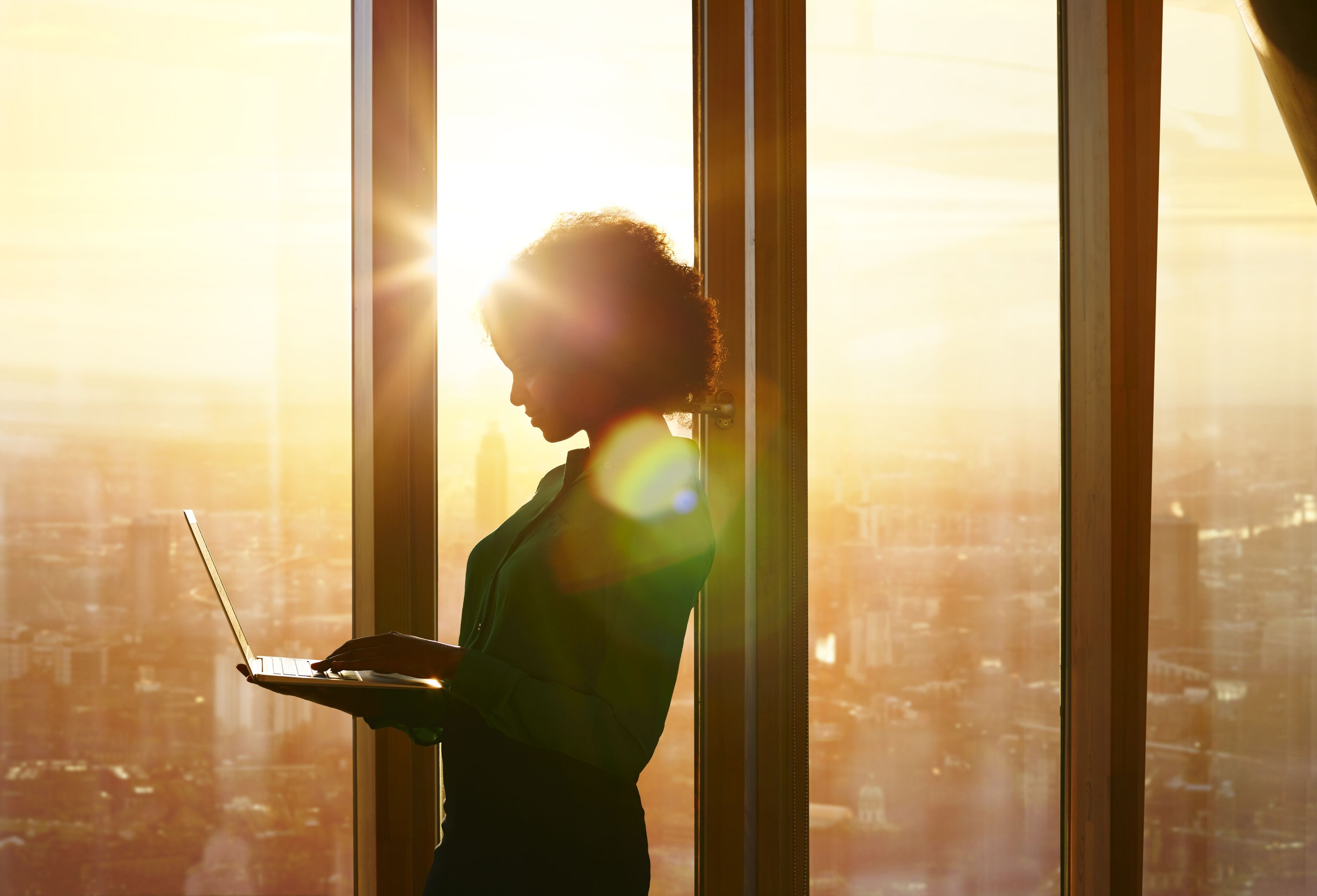 Une femme est debout en silhouette contre une fenêtre, un ordinateur portable à la main, les teintes dorées de l'aube éclairant la ville derrière elle.