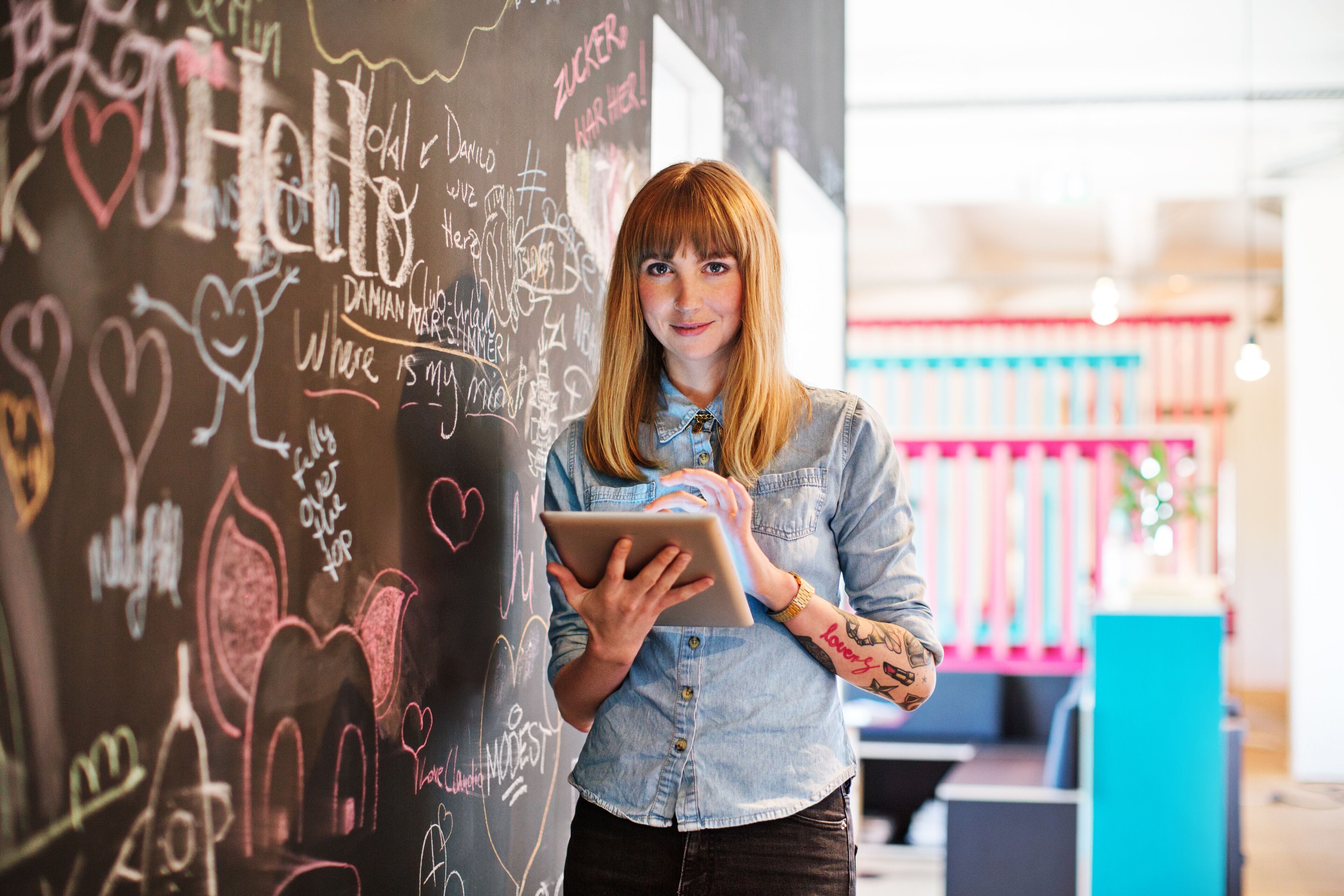 A woman in a denim shirt stands holding a tablet in front of a chalkboard wall filled with doodles and messages.