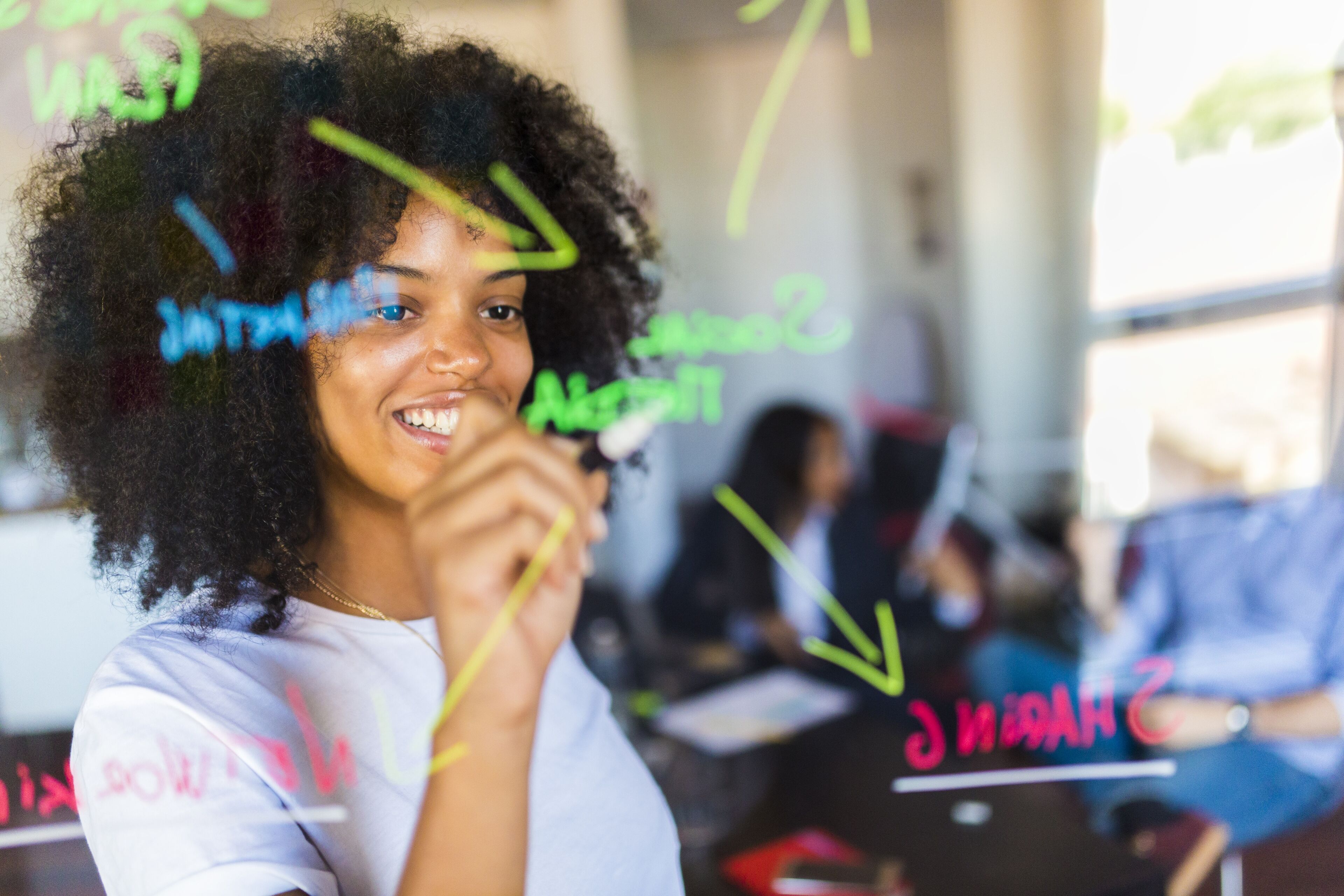 A cheerful young woman writes on a glass wall with markers during a collaborative meeting.