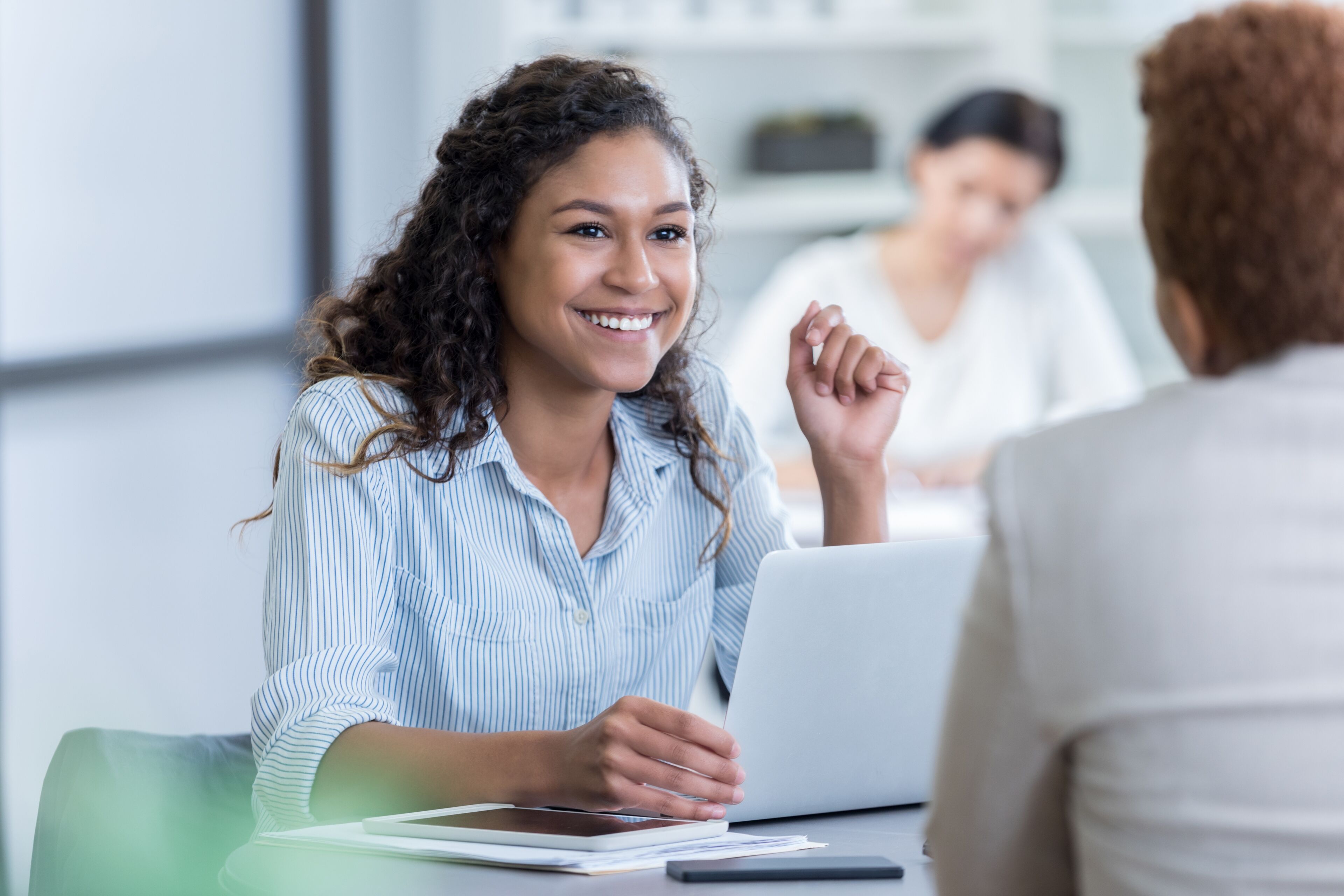 A smiling young woman engaging in a professional discussion with a colleague, with another participant in the background, in a well-lit office setting.