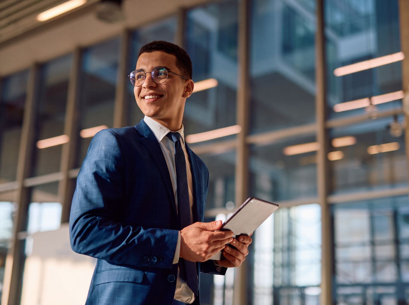 A smiling man wearing glasses and a blue suit, holding a tablet in a modern office setting with large windows.