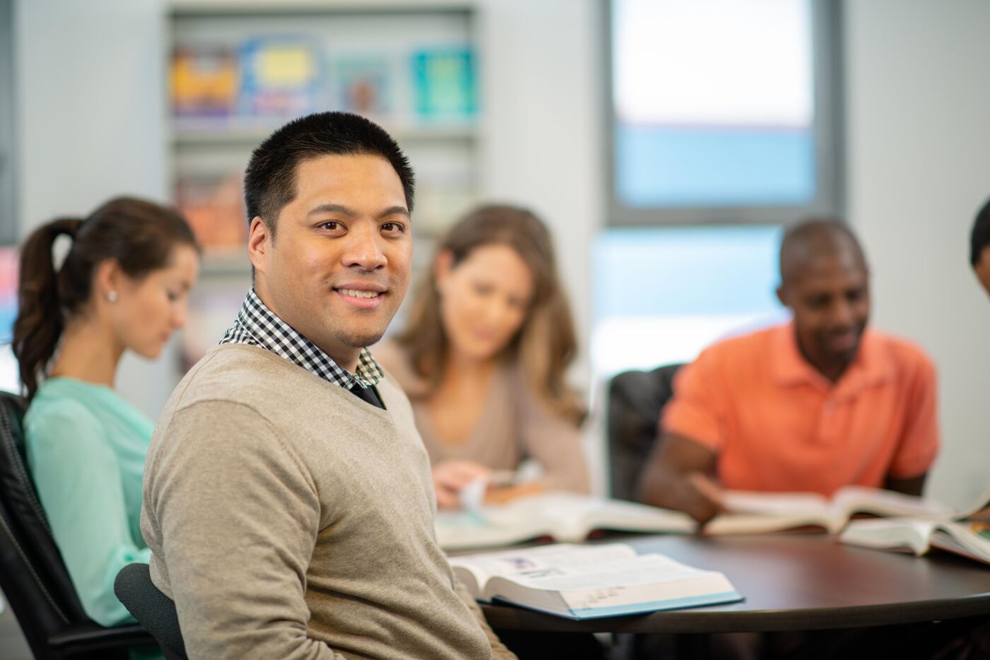 The image shows a group of people sitting around a table, seemingly studying or working together. A man in the foreground is smiling at the camera, while others are focused on books or materials in front of them. The setting appears to be a library or study room.