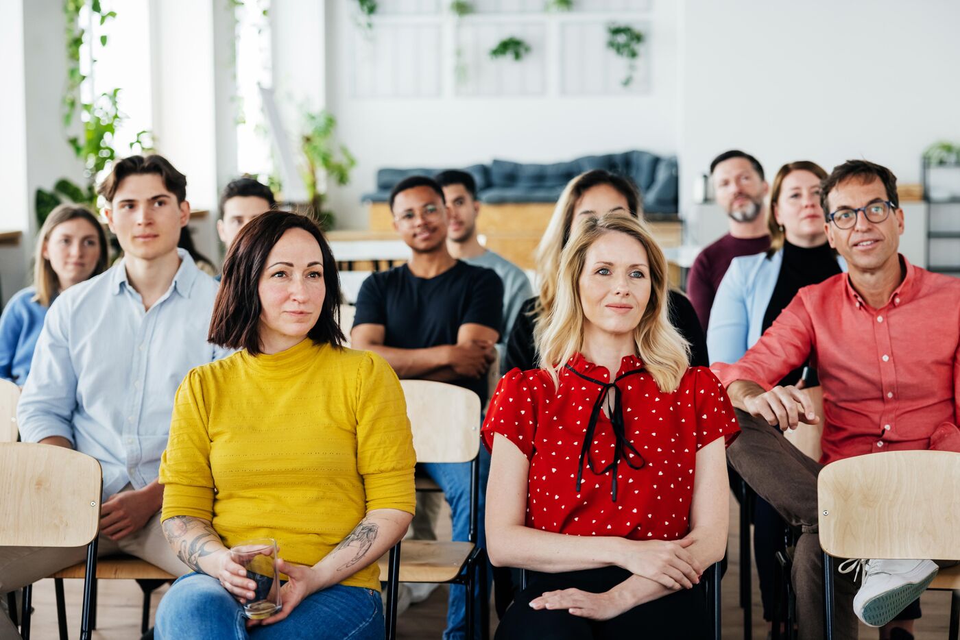 The image shows a group of people sitting in chairs, seemingly at a conference or meeting. They are diverse in age and ethnicity, and appear to be paying attention to a speaker or presentation.