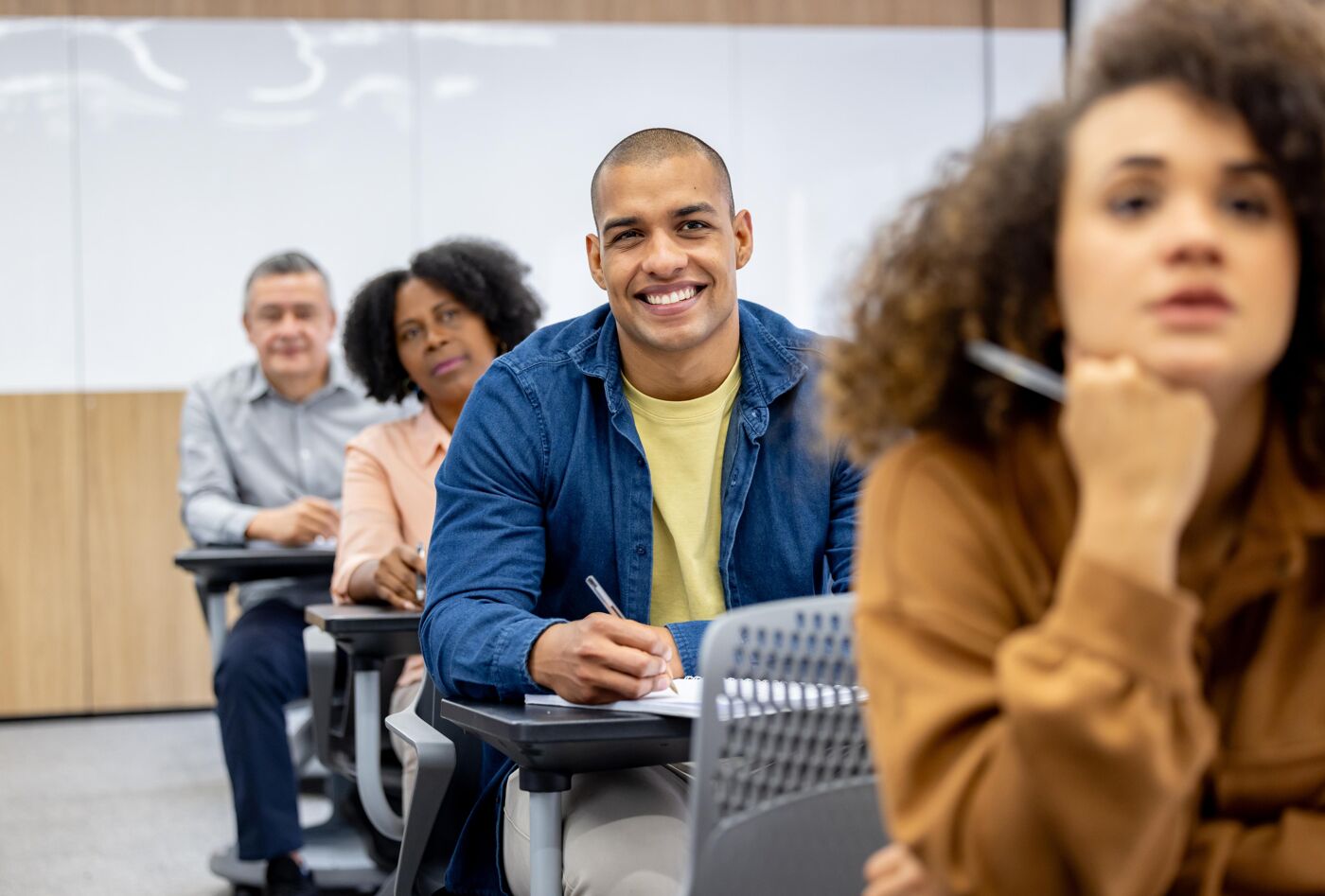 The image shows a diverse group of students in a classroom setting. They are sitting at desks, with some taking notes and others listening attentively. The atmosphere appears to be focused and engaged.