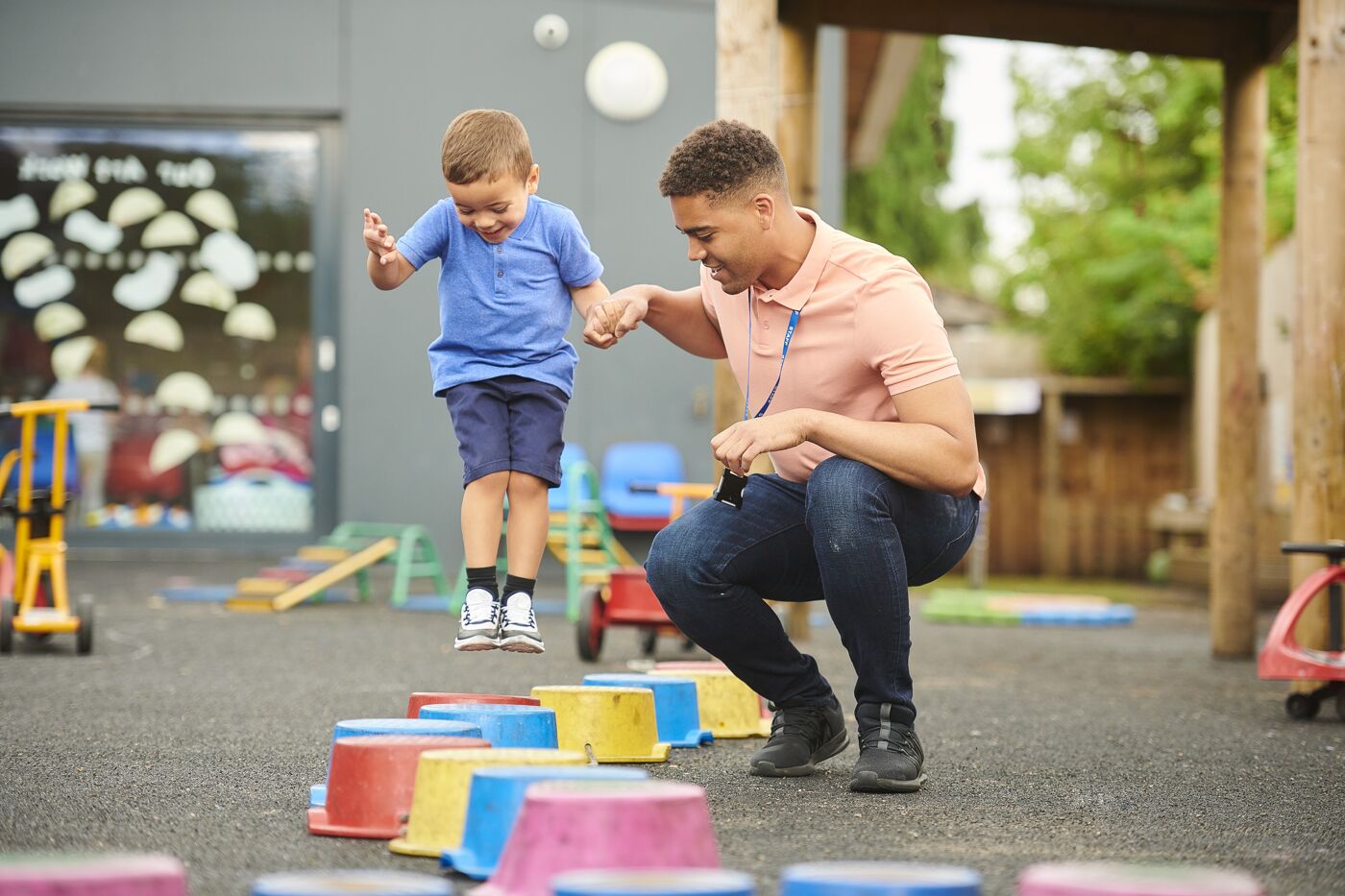 A young boy takes cautious steps across a balance path with the support of a daycare worker, outdoors in a play area.