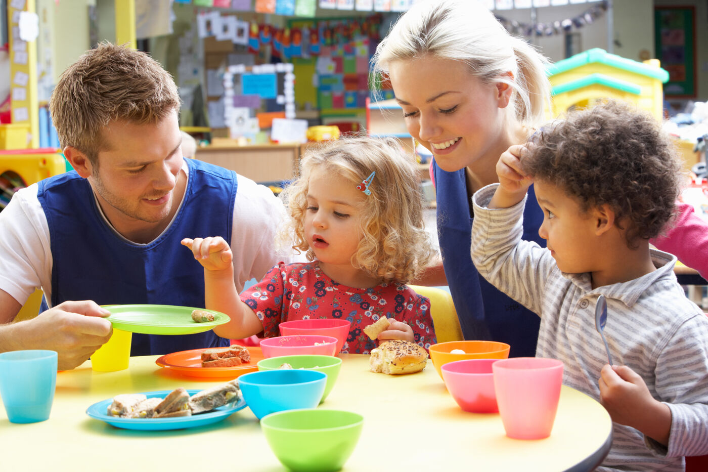 Two daycare workers assist young children with their meals at a colorful table, fostering a cheerful and nurturing environment.