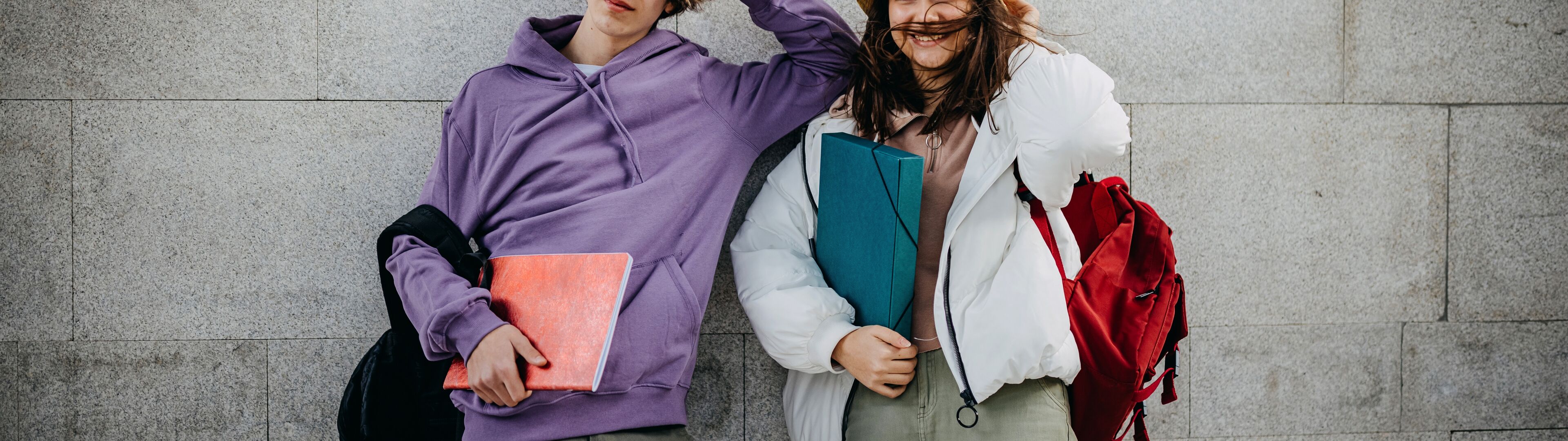 Two cheerful students in casual attire pose against a grey wall, each holding a notebook.