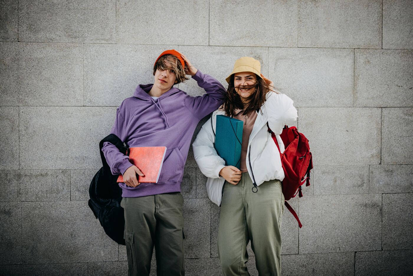 Two cheerful students in casual attire pose against a grey wall, each holding a notebook.