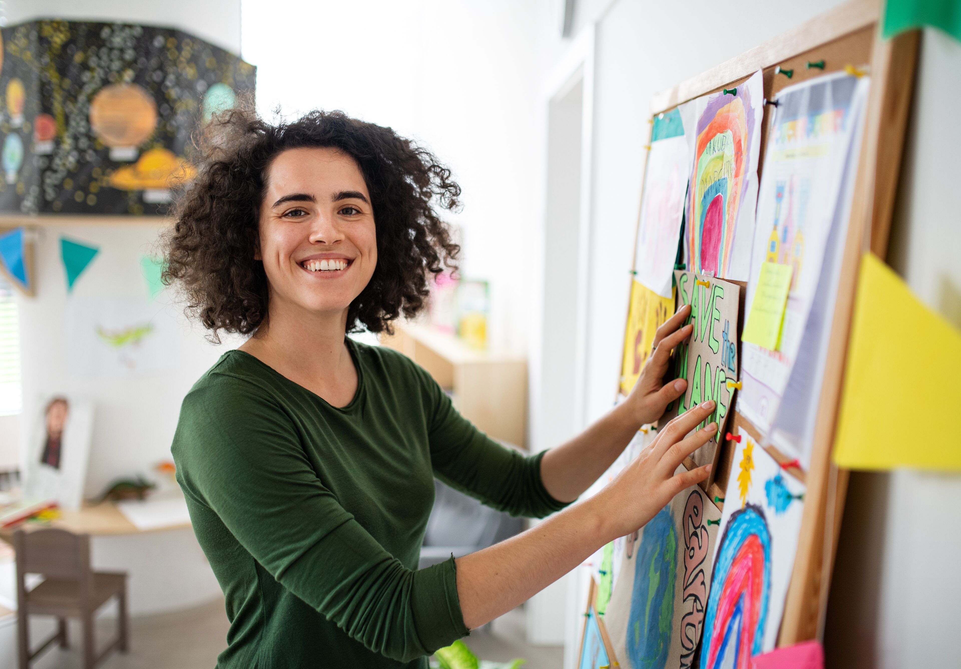 A cheerful art teacher arranges colorful student artwork on a display board in a bright classroom.