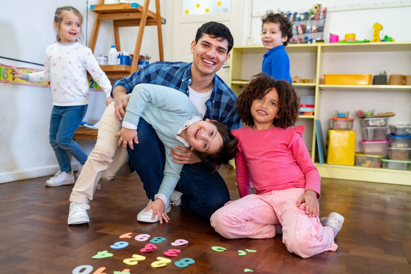 A daycare teacher interacts playfully with children in a lively playroom, highlighting a joyful and active childcare environment.