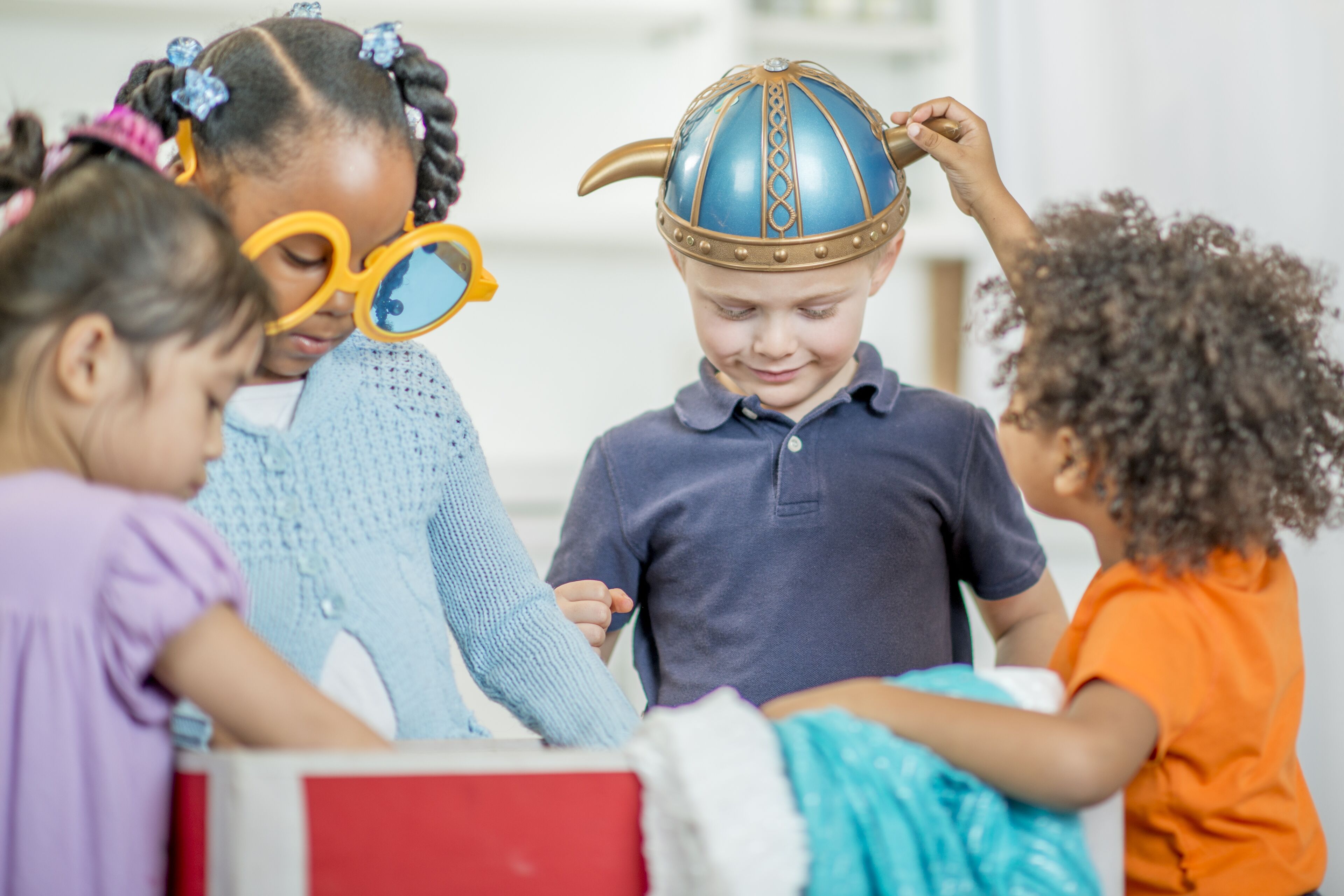 A group of four children are engaged in a playful dress-up activity, with one boy smiling while a girl places a Viking helmet on his head.