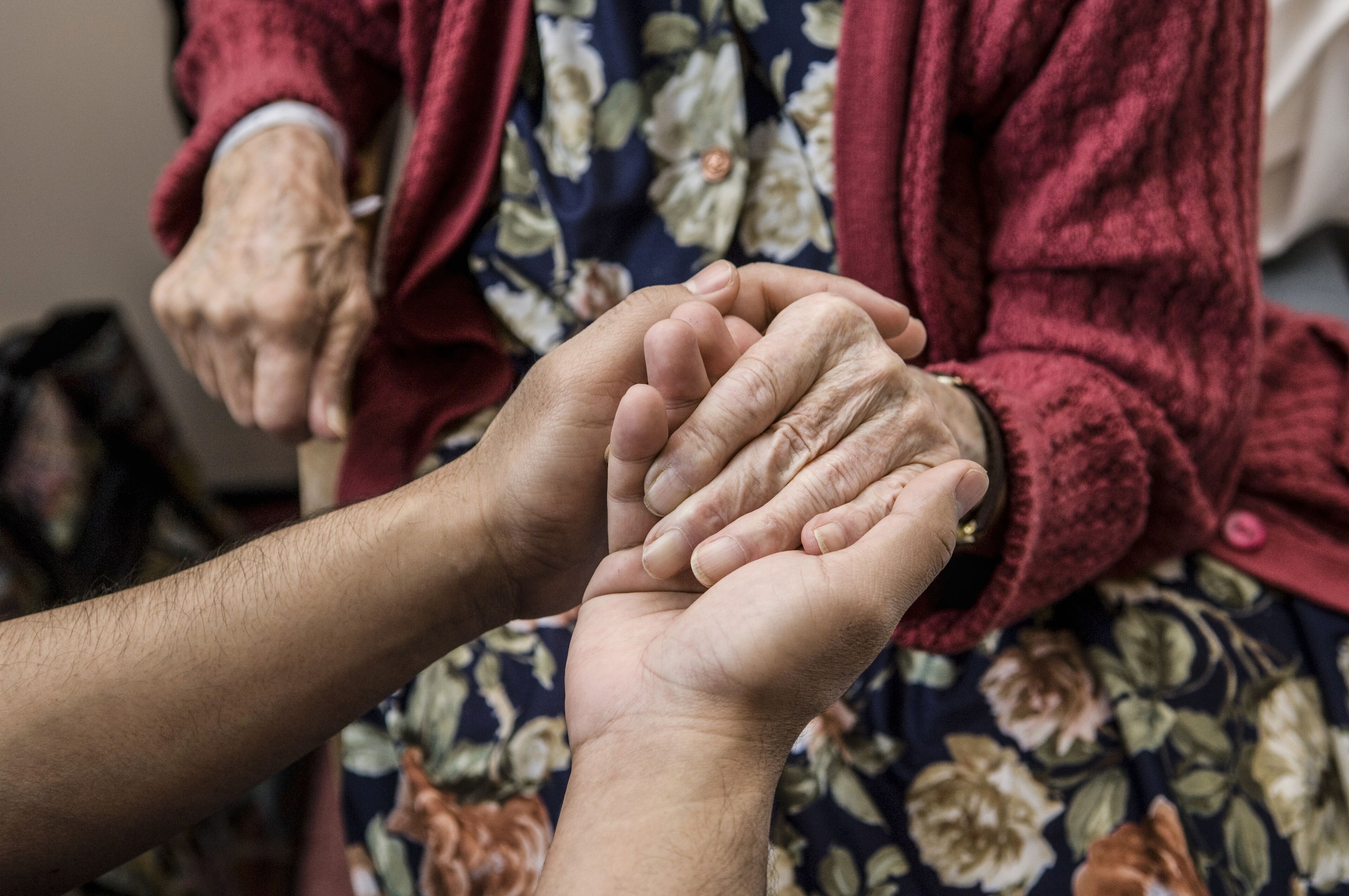 Une personne âgée en pull rouge tenant la main d'un plus jeune, symbolisant le soin et la connexion.
