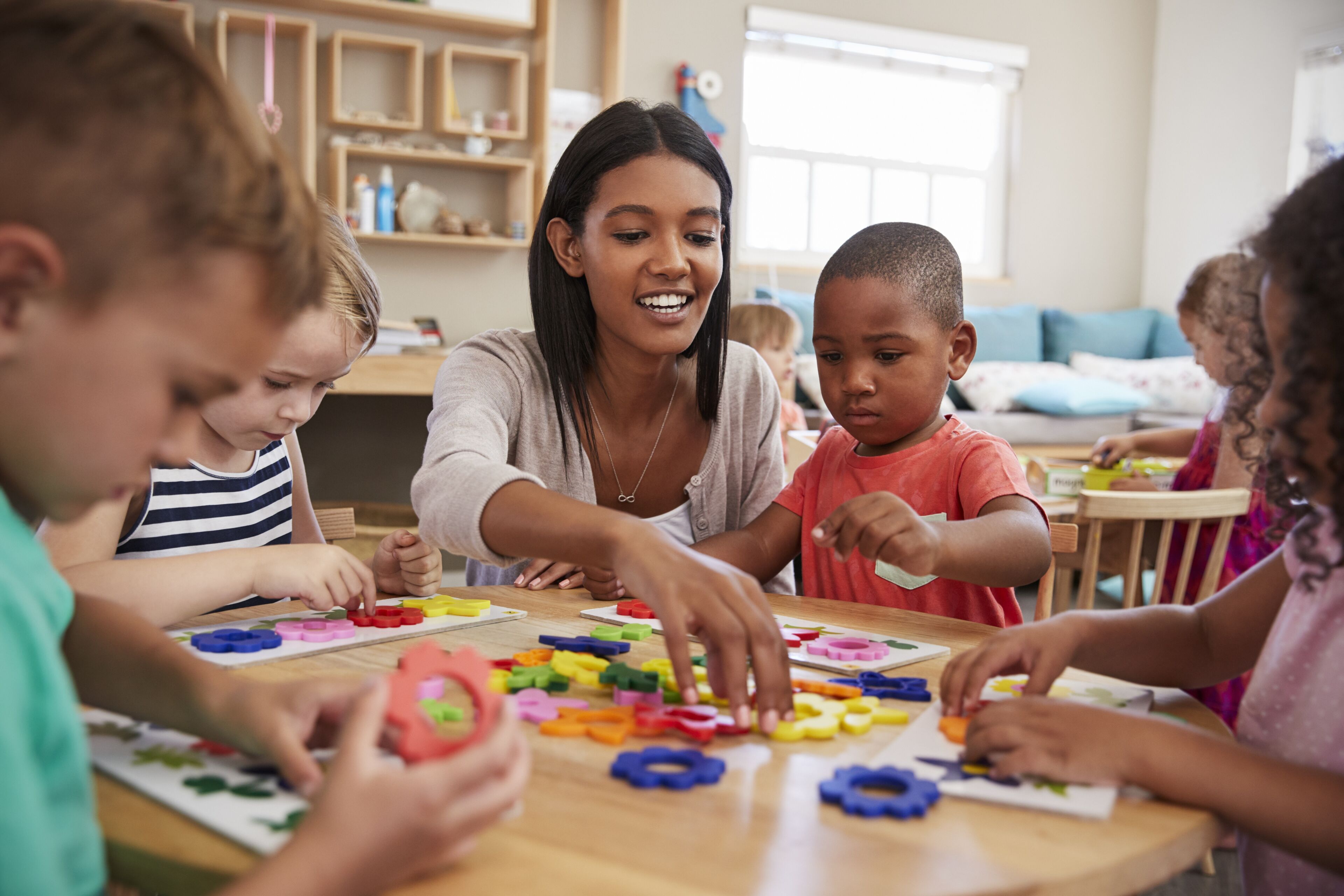 Une enseignante interagit avec de jeunes enfants autour d'une table, les guidant dans une leçon ludique sur les formes et les couleurs à l'aide de jouets éducatifs.