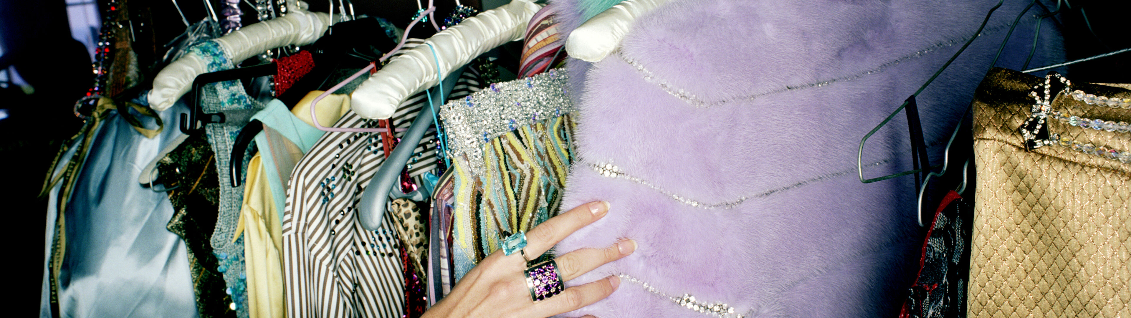 A hand touches a furry purple garment among a variety of colorful clothes on hangers.