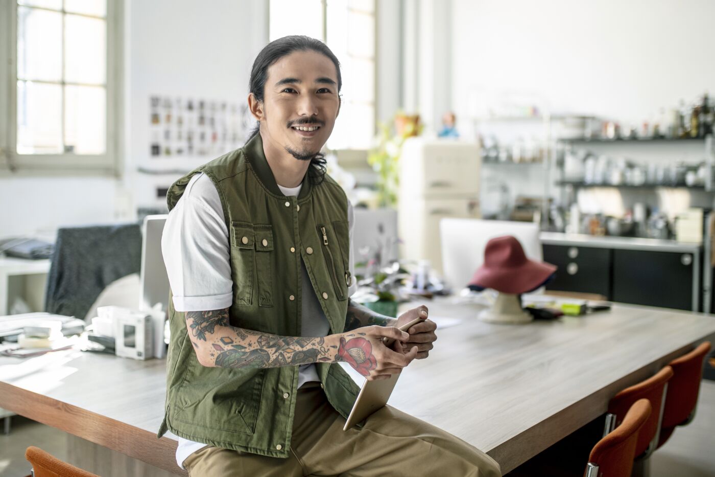 A smiling tattooed man sits at a wooden table in a well-lit creative studio.