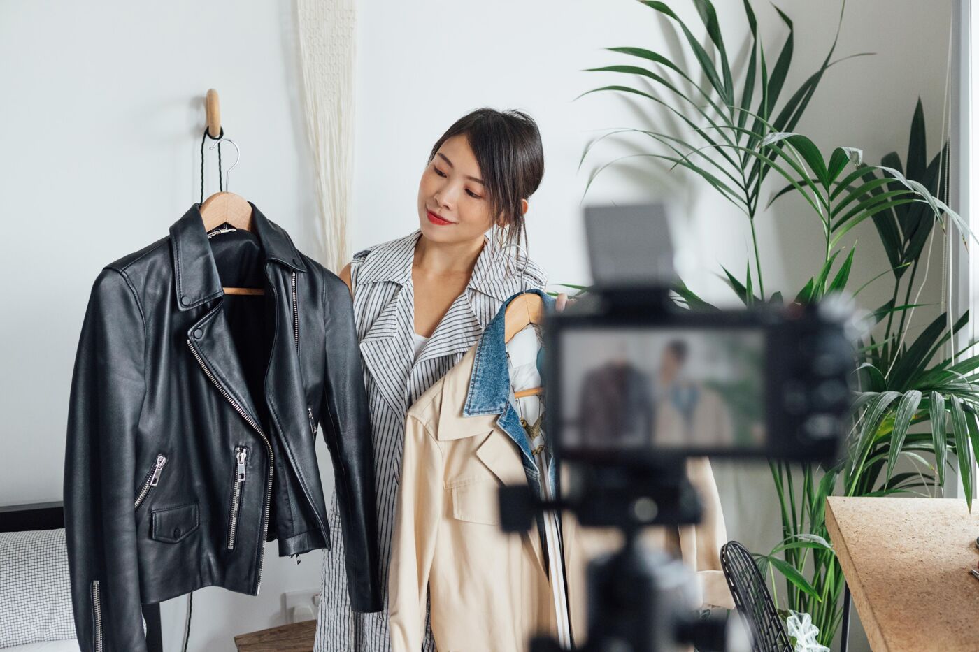 A young woman stands in front of a camera, holding up a black leather jacket and other clothing items for a fashion video.
