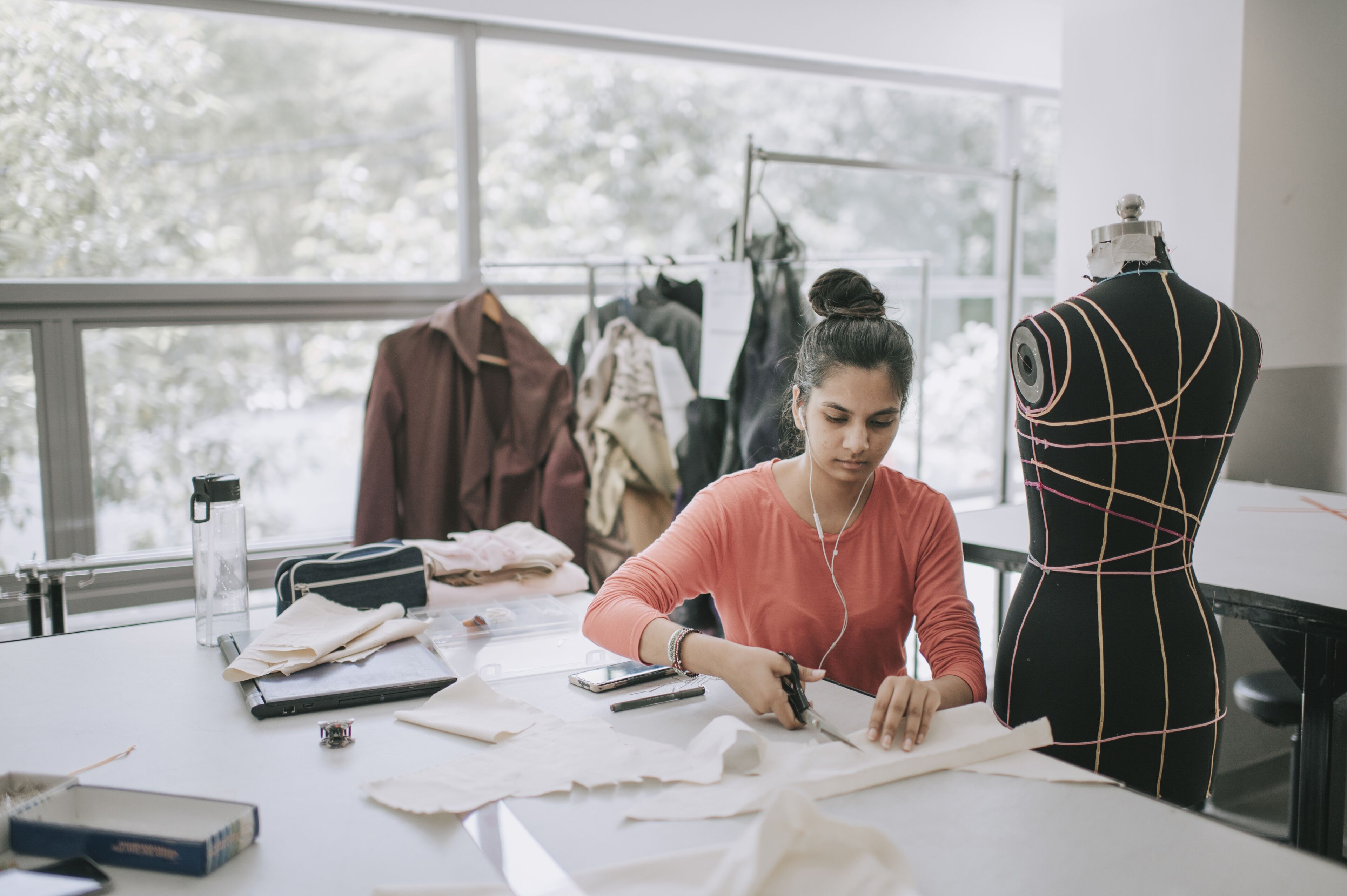 Une créatrice de mode concentrée coupe du tissu à côté d'un mannequin, avec des vêtements et une machine à coudre en arrière-plan.