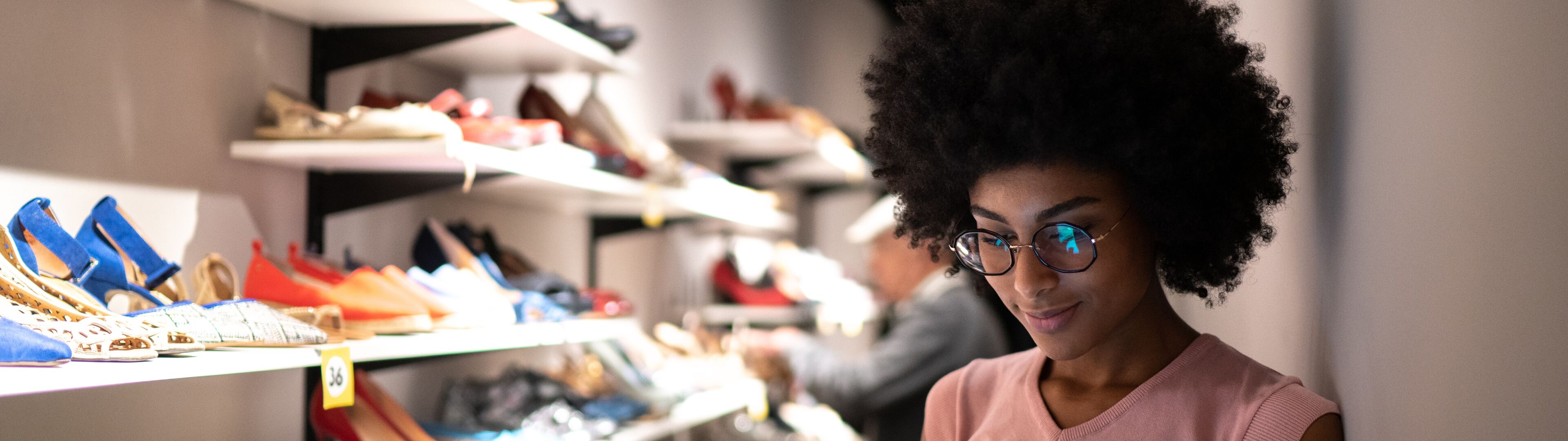 A woman with an afro hairstyle examines a tablet in front of a display of colorful shoes in a store.