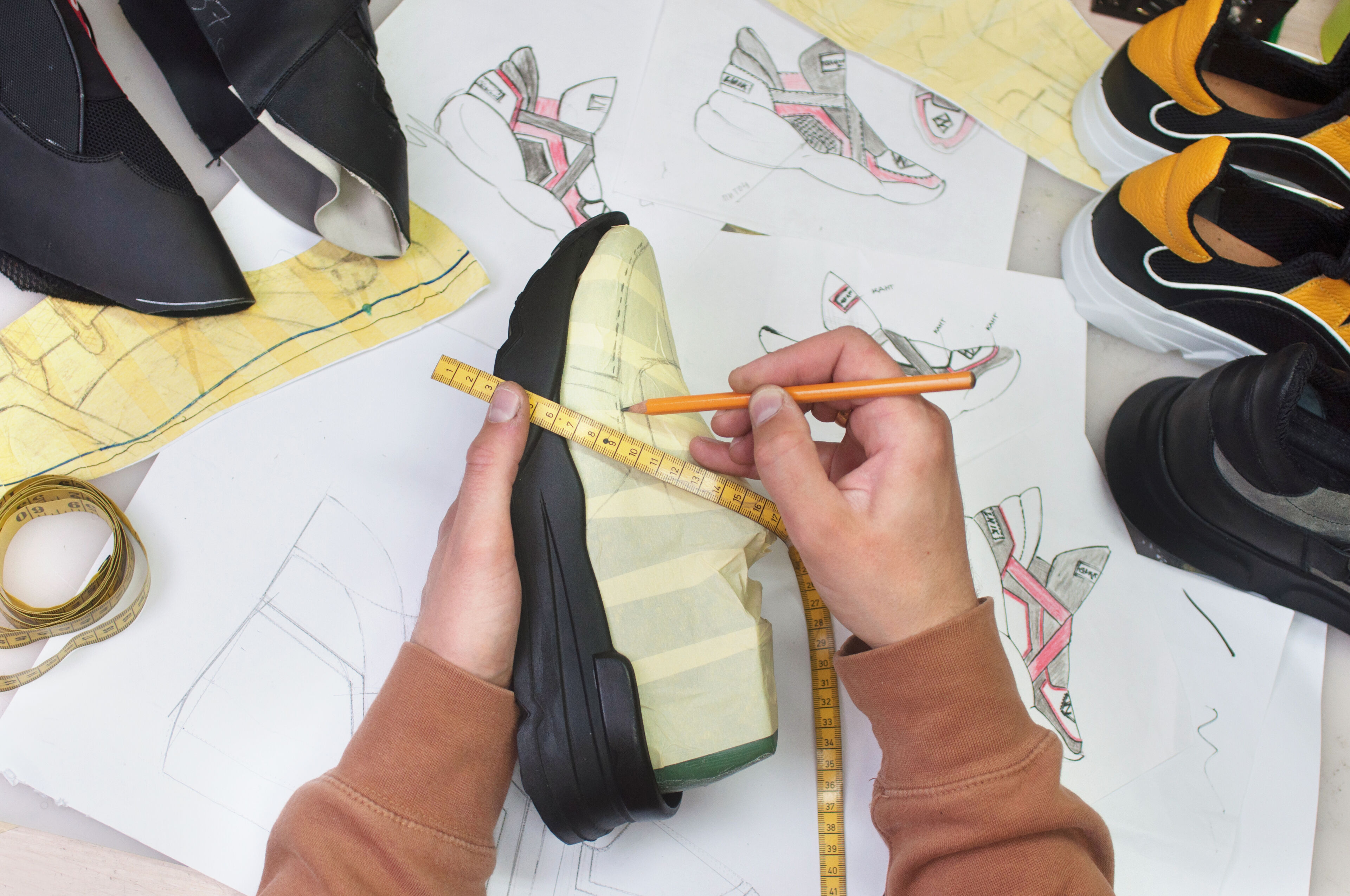 A hand measures a sneaker prototype with a tape measure amid sketches and other prototypes.