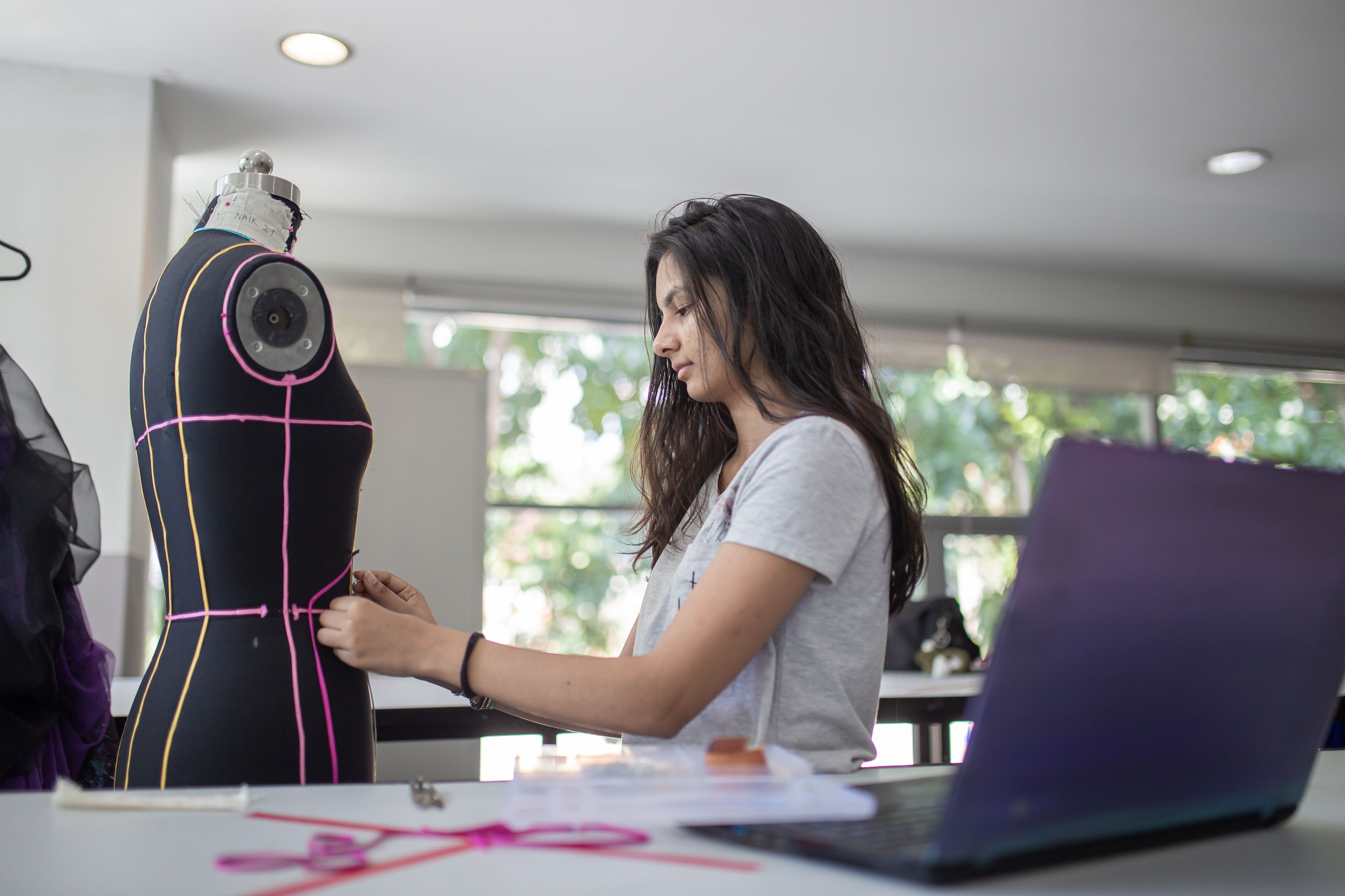 Une créatrice de mode concentrée ajuste un vêtement sur un mannequin à côté de son ordinateur portable dans un atelier bien éclairé.